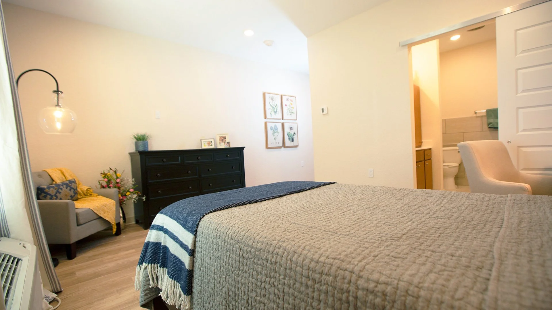 A cozy bedroom with a large bed covered in a beige quilt and a blue and white striped throw blanket. To the left is a sitting area with a gray armchair decorated with pillows and a yellow throw, accompanied by a floral arrangement. A black dresser wi