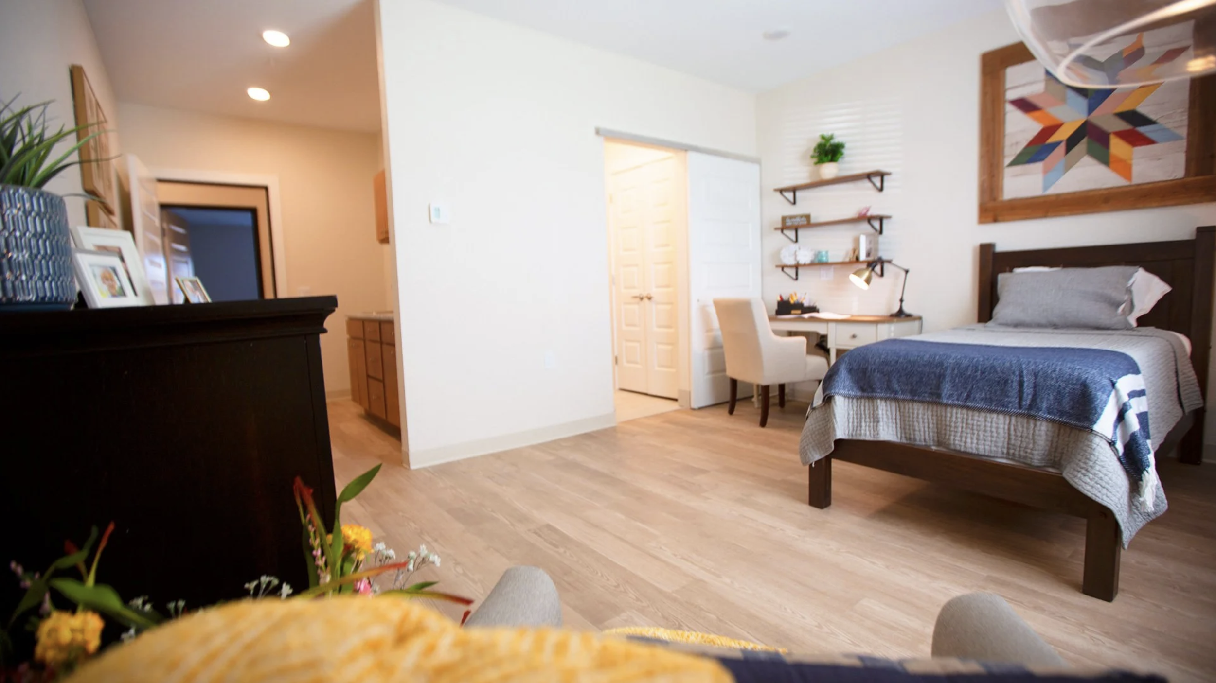 A cozy bedroom with a bed that has a wooden headboard, a gray and blue striped blanket, and a matching pillow. There are floating shelves and artwork on the wall, a small writing desk with a beige chair, and a lush potted plant on a dresser. The floo
