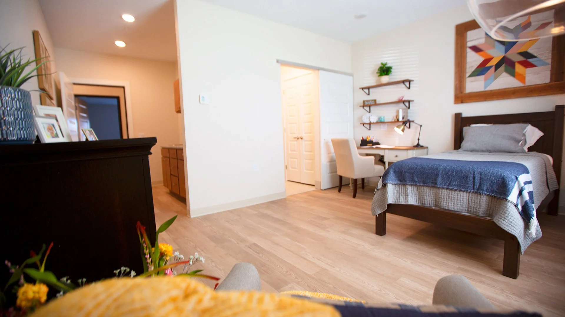 A cozy bedroom with a bed that has a wooden headboard, a gray and blue striped blanket, and a matching pillow. There are floating shelves and artwork on the wall, a small writing desk with a beige chair, and a lush potted plant on a dresser. The floo
