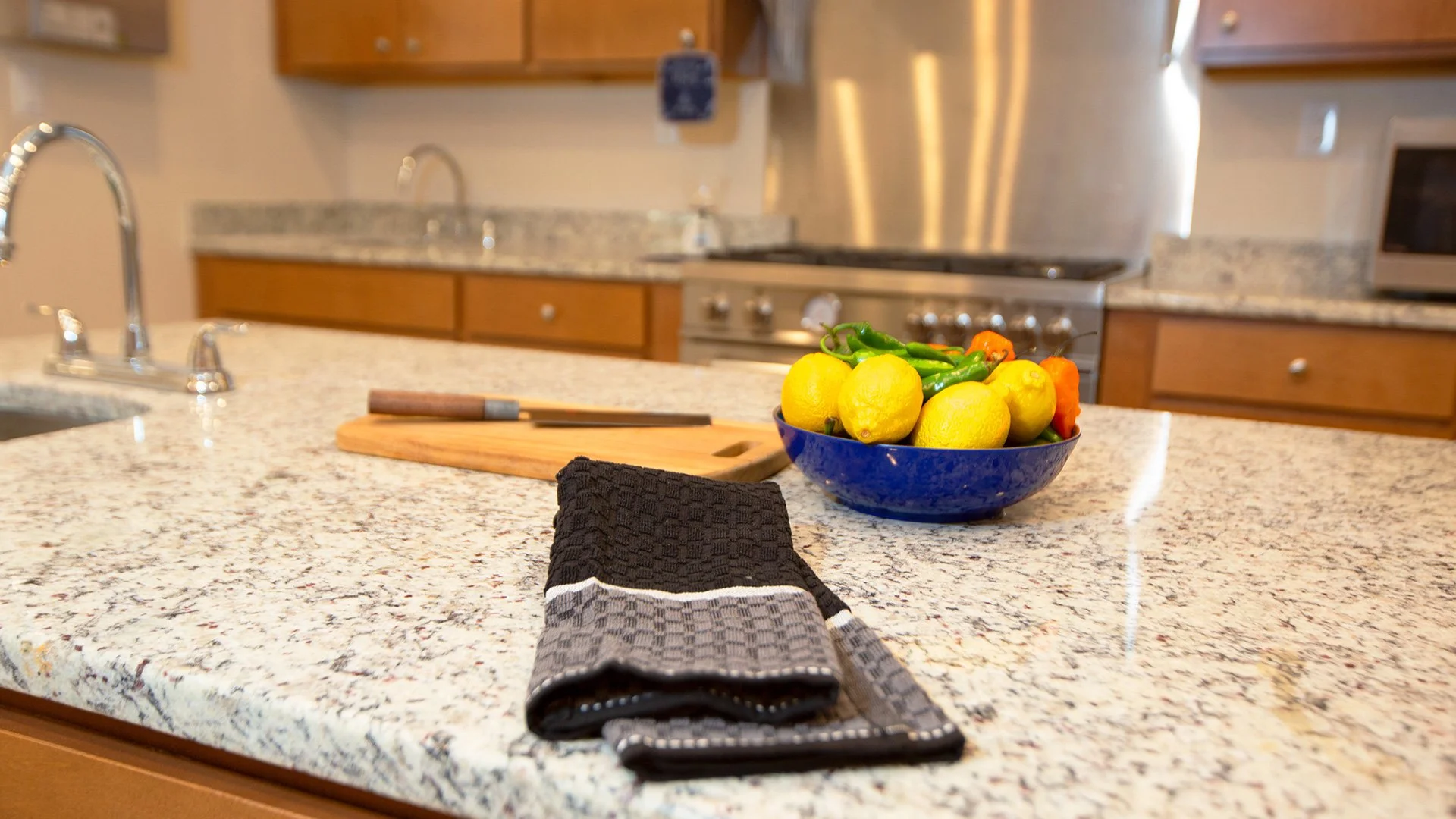 Kitchen countertop with a bowl of lemons and peppers, a wooden cutting board, a black kitchen towel, and a knife, with a stove and cabinets in the background.