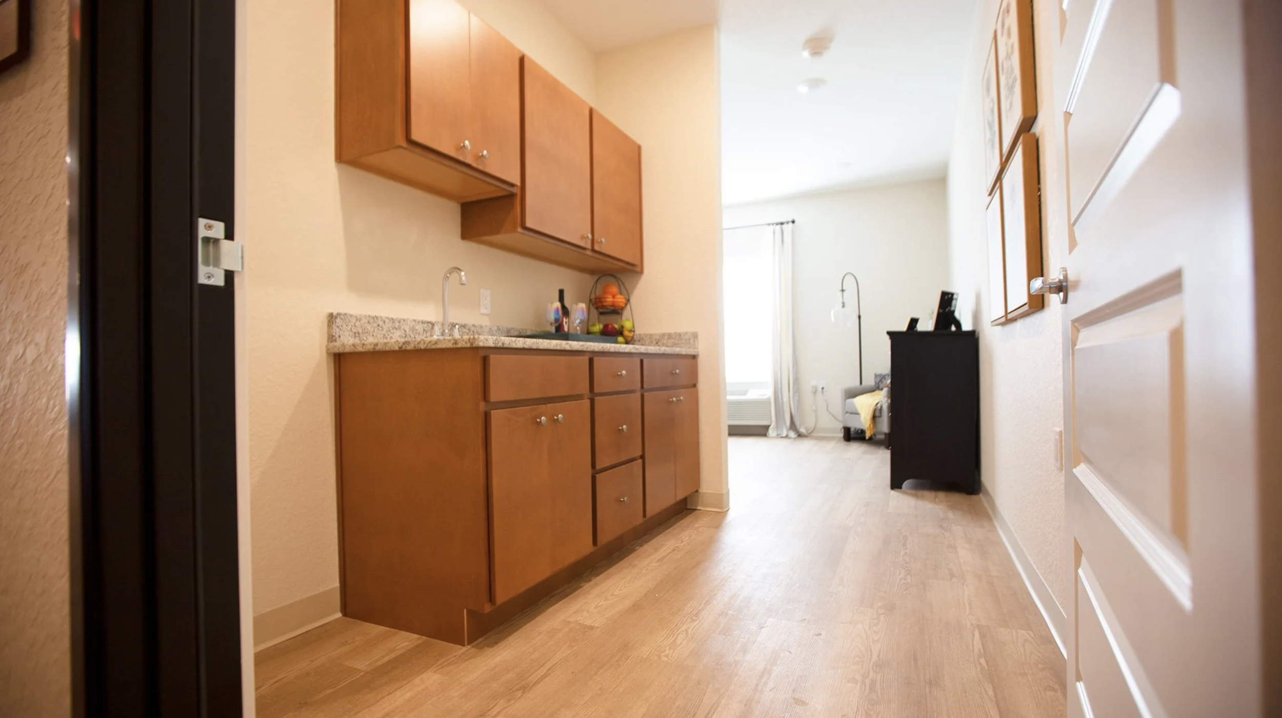 Interior view of a small apartment with a kitchen area featuring wooden cabinets, a granite countertop, and a sink. Past the kitchen, a living area is visible with a window, a radiator, and some furniture, including a black dresser or cabinet and a s