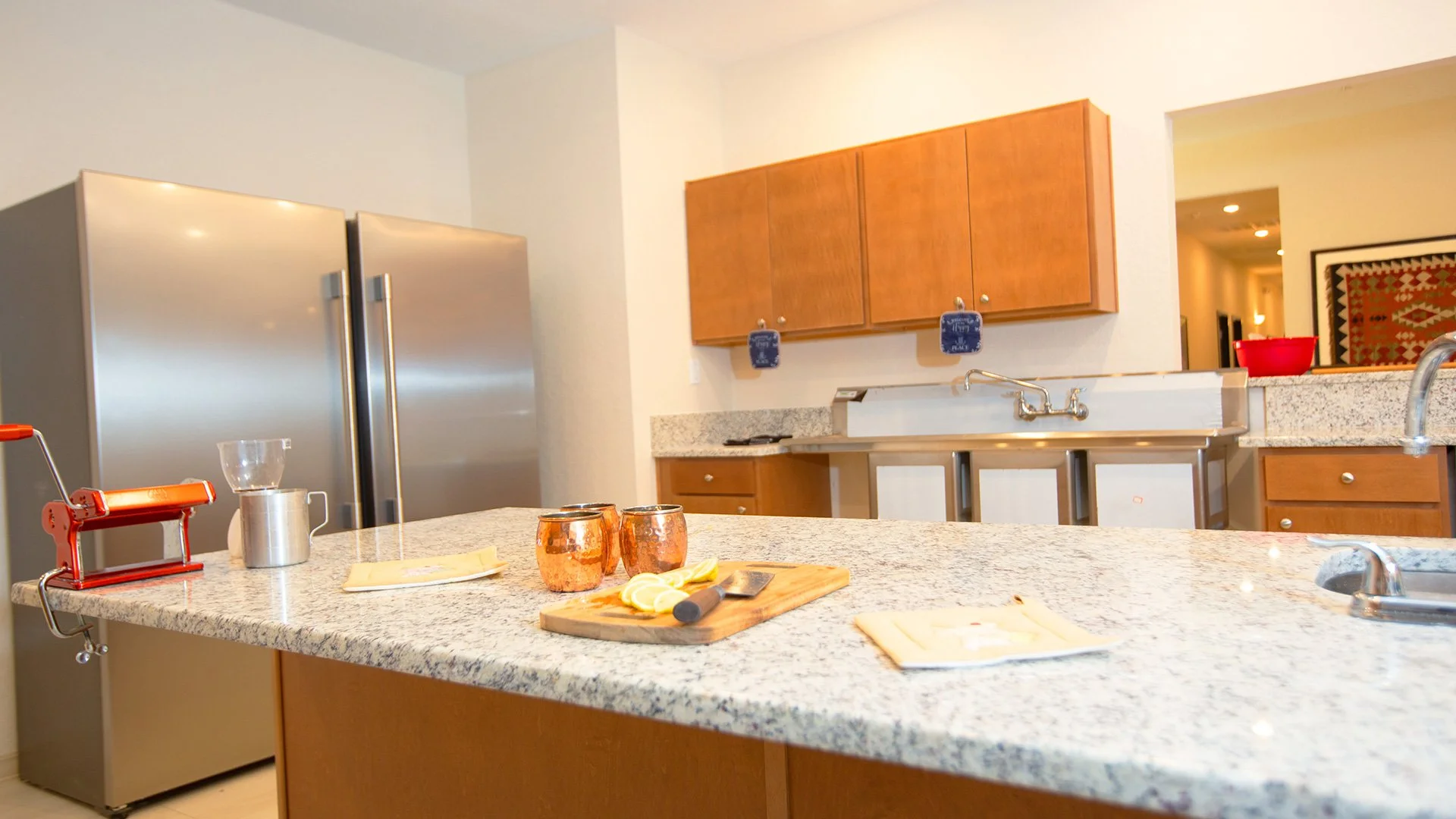 Kitchen with granite countertops, wooden cabinets, a stainless steel refrigerator, a double sink, and a kitchen island with lemons, copper mugs, and a pizza cutter. A red manual pasta machine is on the island.