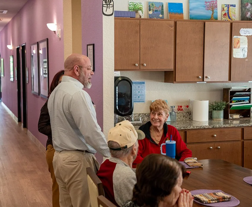 A group of elderly people and a middle-aged man gathered around a kitchen counter, engaged in a friendly conversation in a community or senior center setting.