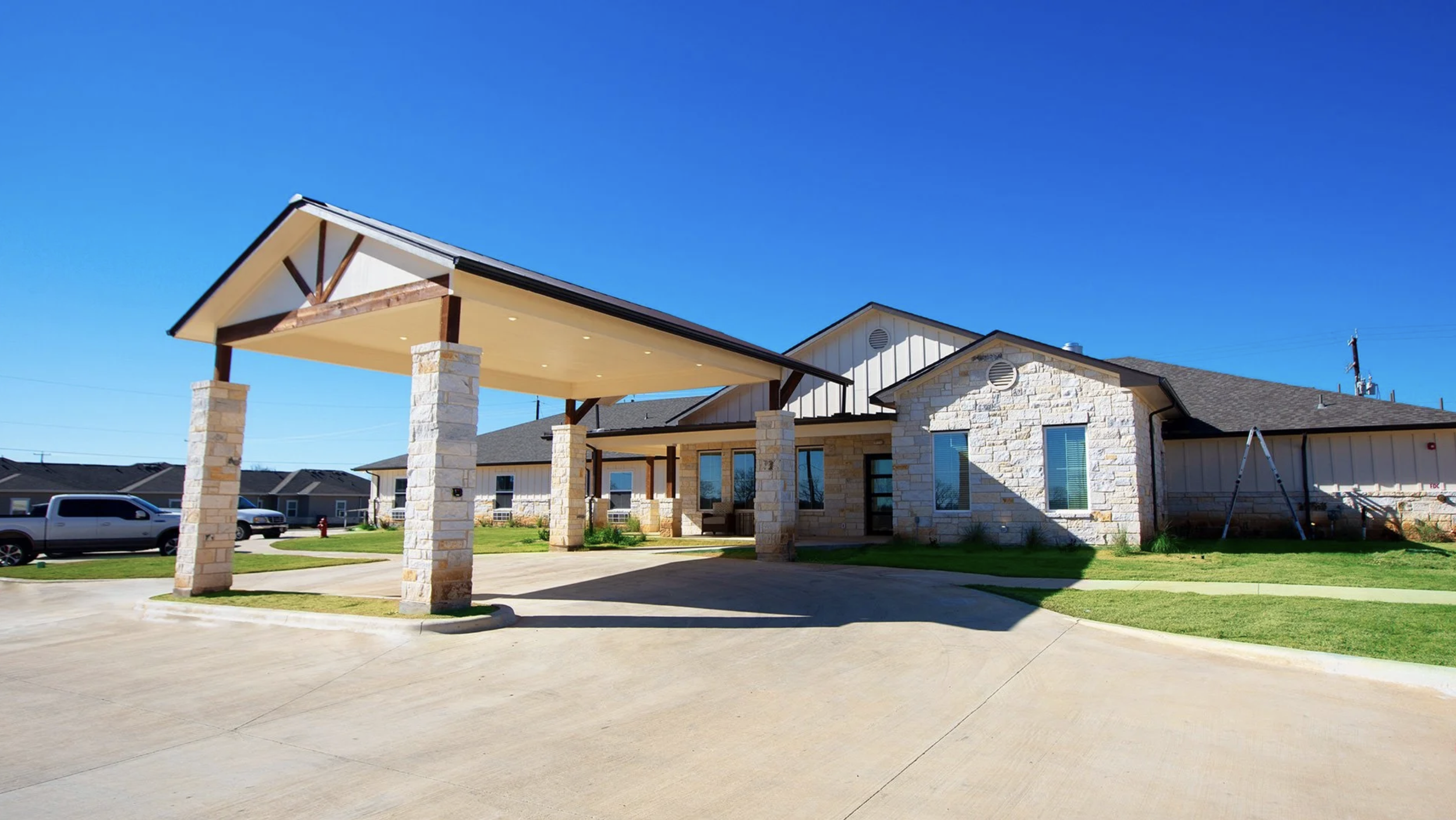 Modern residential building with stone and siding exterior, covered entrance with stone columns, parked vehicles, and clear blue sky.