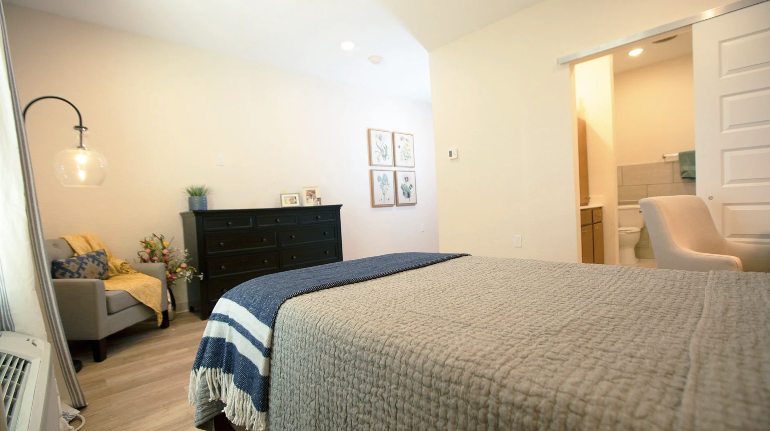 A cozy bedroom with a large bed covered in a beige quilt and a blue and white striped throw blanket. To the left is a sitting area with a gray armchair decorated with pillows and a yellow throw, accompanied by a floral arrangement. A black dresser wi