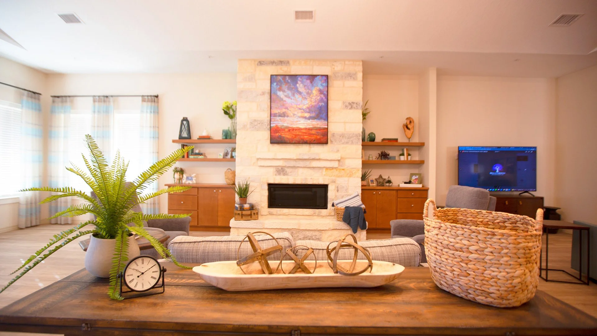 Living room with a central stone fireplace, some wall-mounted wooden shelves, a television, and a variety of decorative items. Potted plants and a dining table with a clock and decorative objects in the foreground.