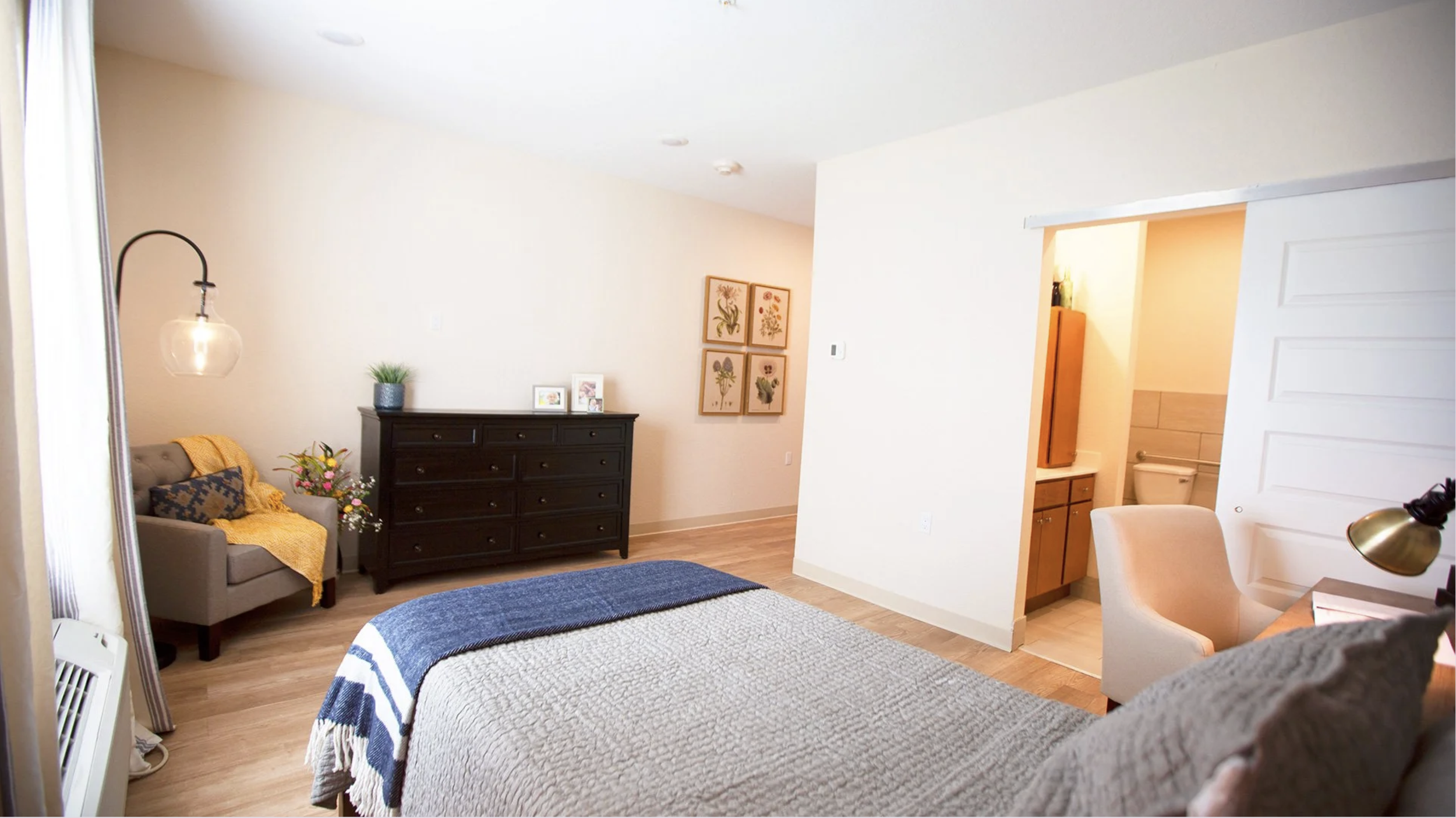 A cozy bedroom with a bed in the foreground, a gray armchair with a yellow blanket and pillows, a white lamp, a black dresser with framed pictures and a potted plant, wall art with botanical prints, an adjacent bathroom with a visible toilet, and a w