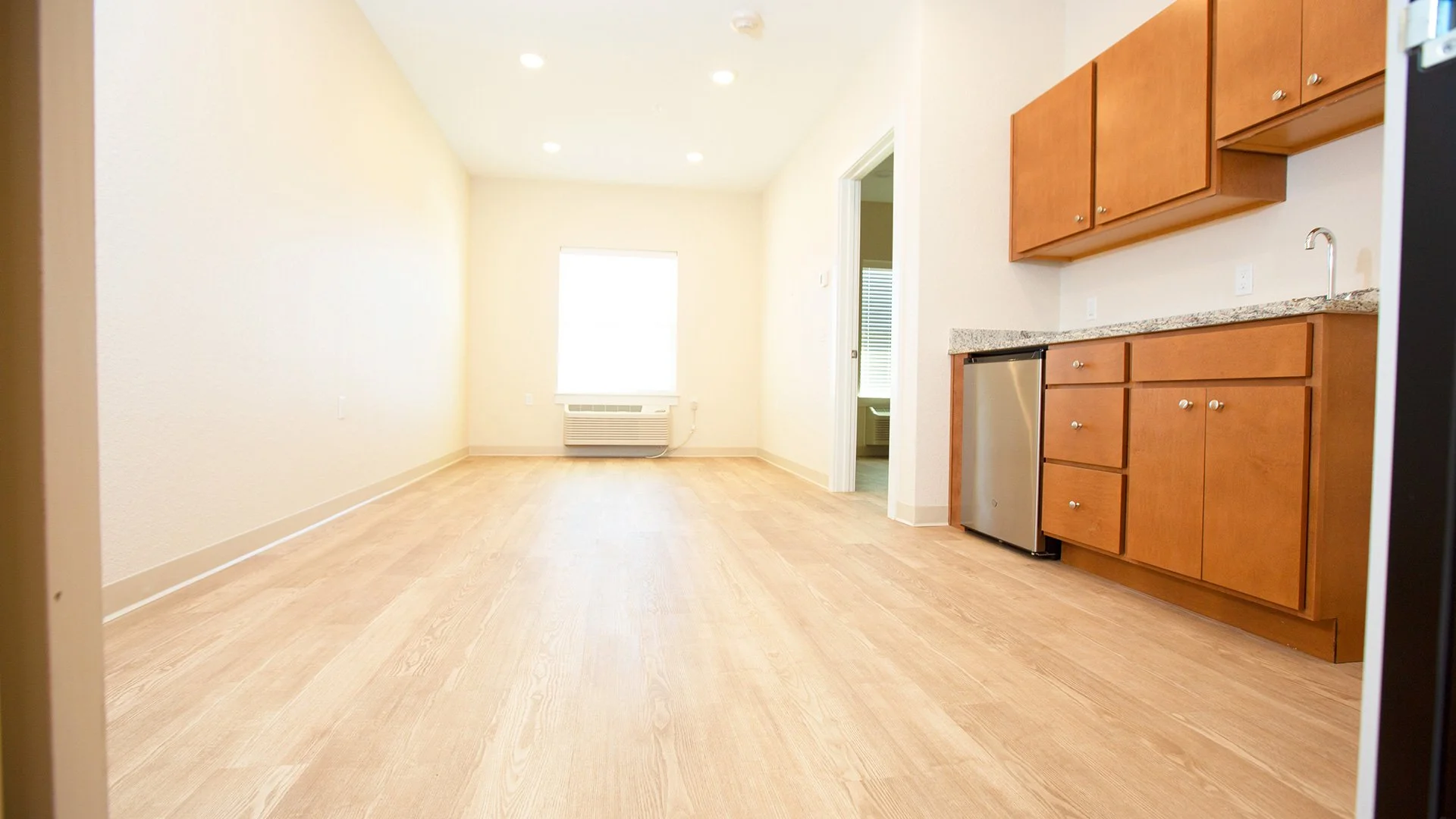Empty room with light-colored wooden floors, beige walls, and large window with blinds. Kitchen area on the right side with brown cabinets, granite countertop, a small refrigerator, and a sink. Open doorway to another room.