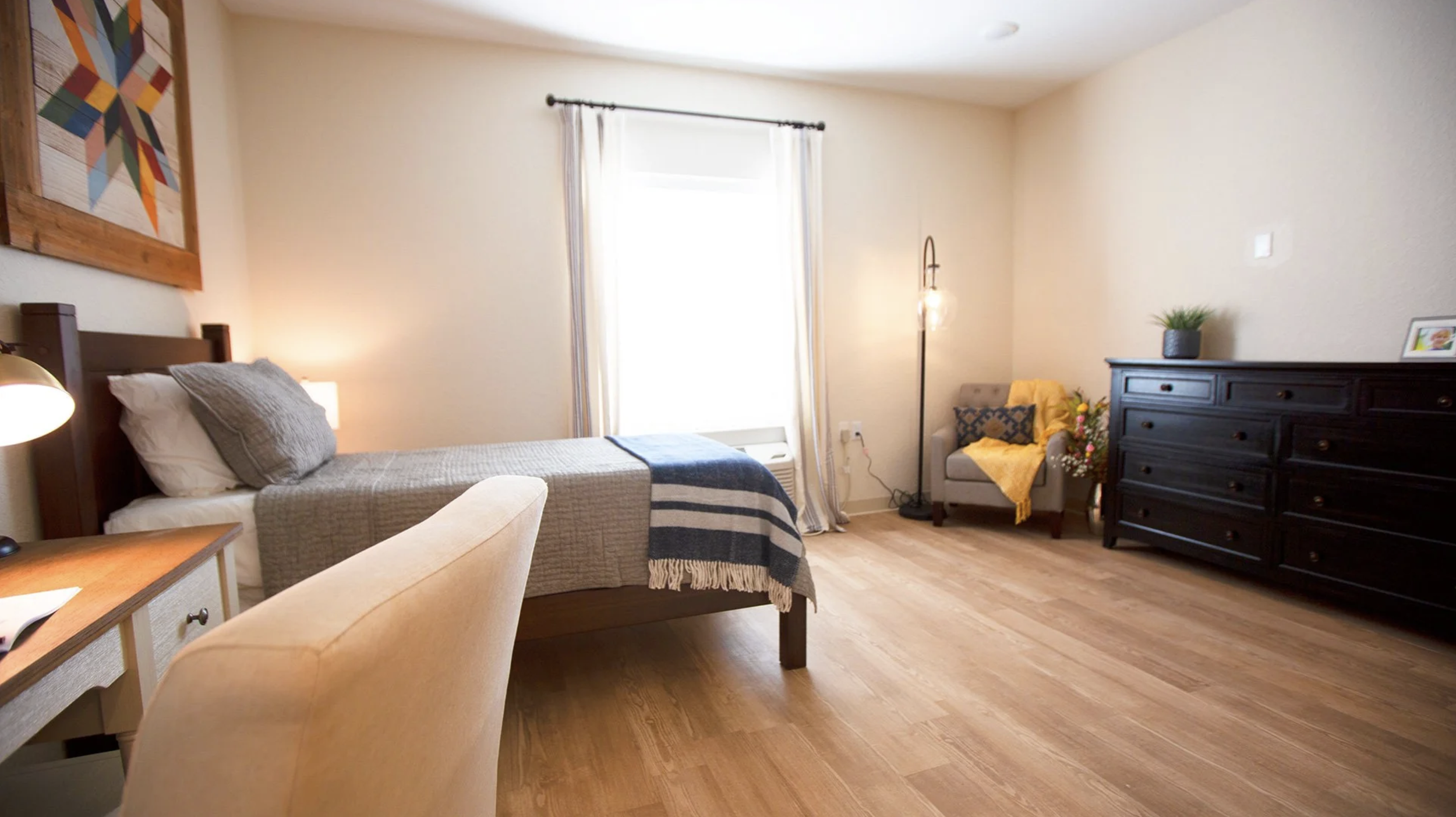 A bedroom with a bed, nightstand, armchair, dresser, and a window with curtains, decorated with plants and artwork.