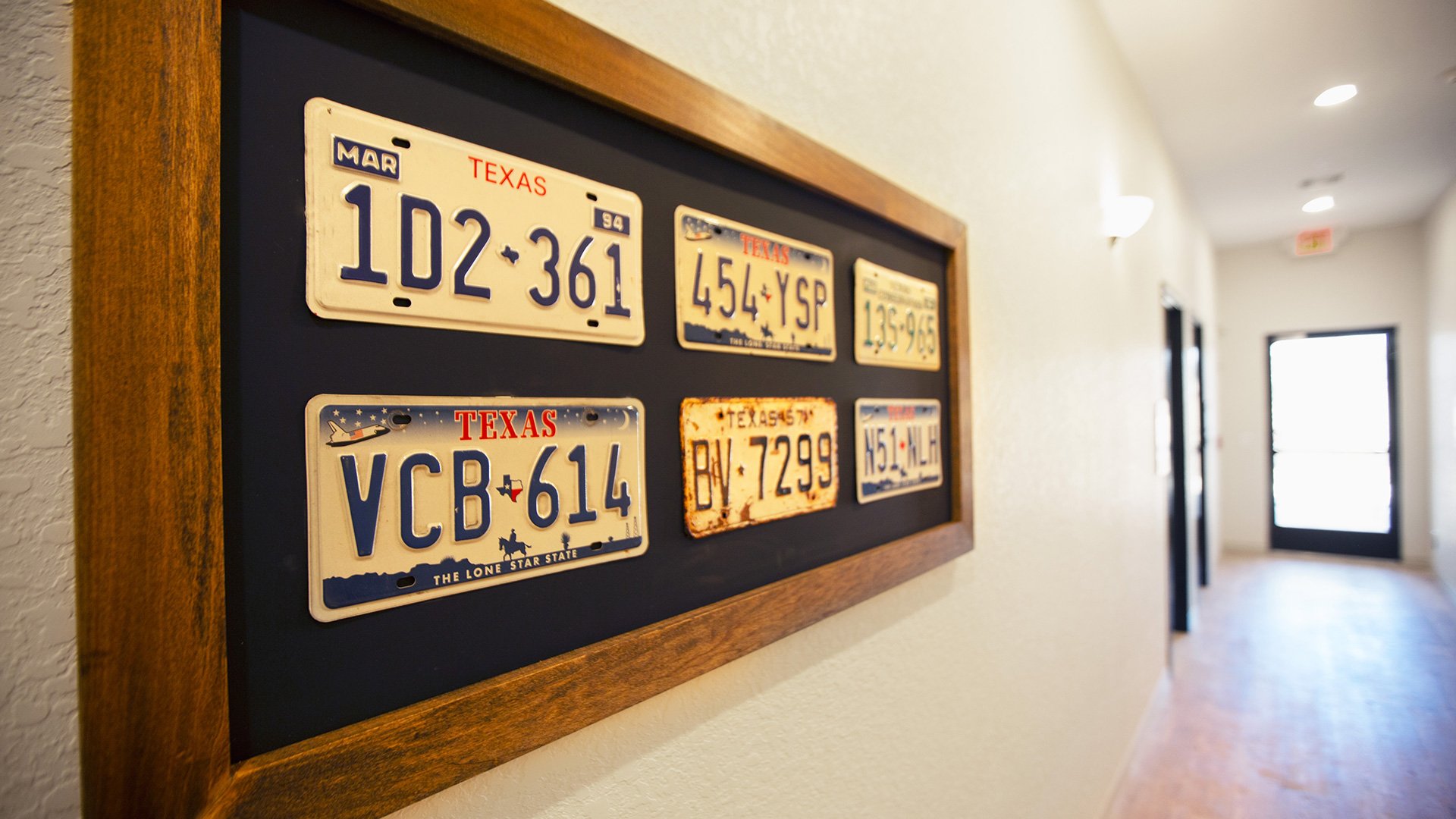 A framed display of vintage Texas license plates on a black background, hung on a beige wall in a hallway.