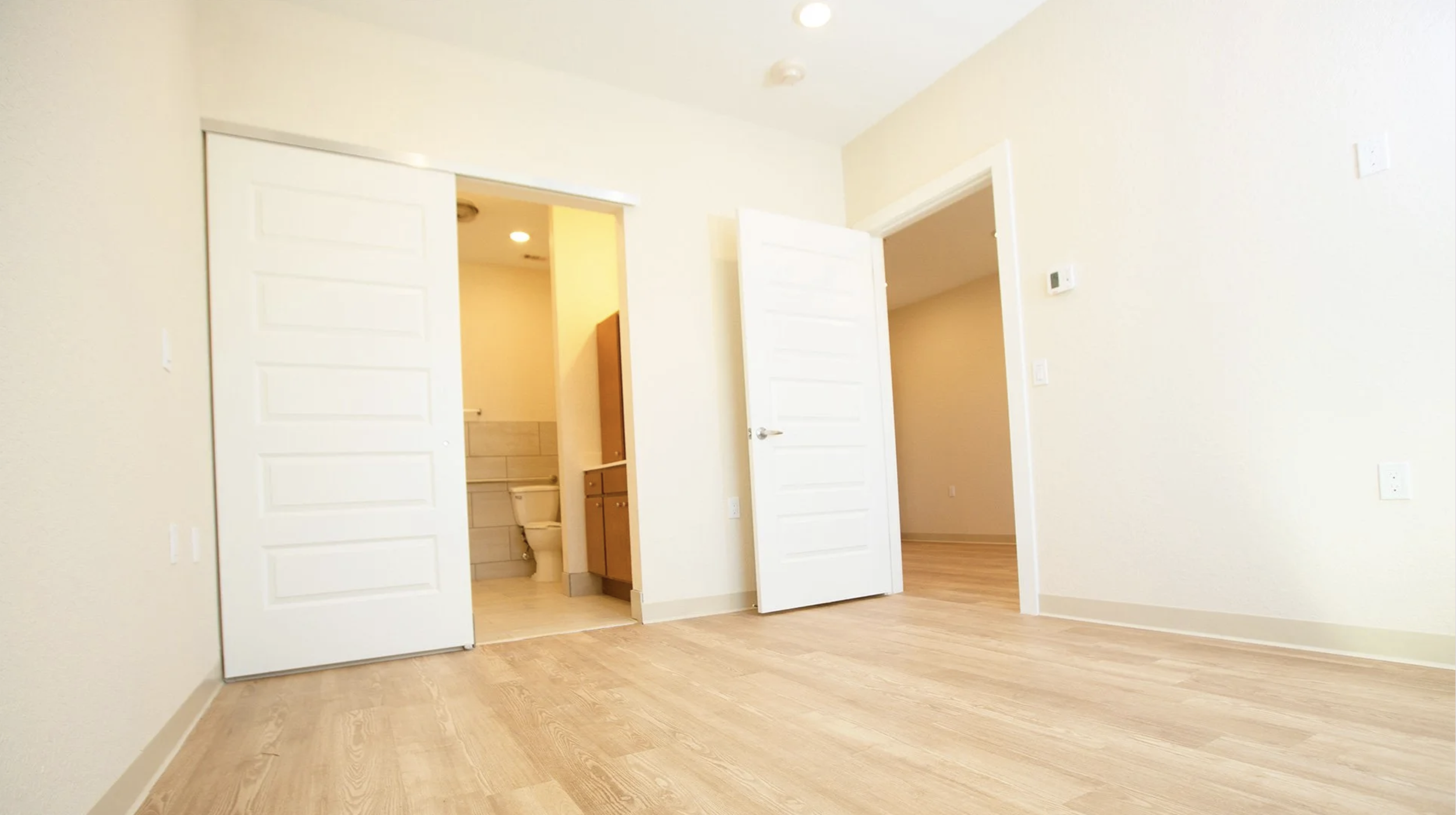 Empty room with light-colored walls, wooden floor, two open doors leading to a bathroom and another room, and visible electrical outlets and thermostat on the walls.