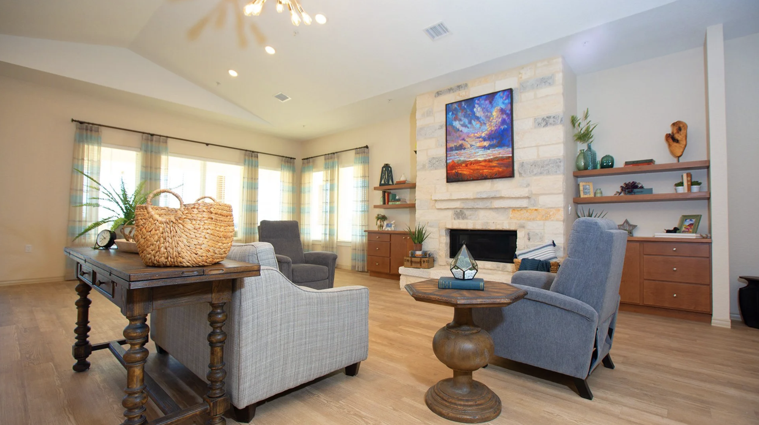 Living room with a stone fireplace, colorful painting above it, wooden and upholstered furniture, decorative plants, and a large window with striped curtains.