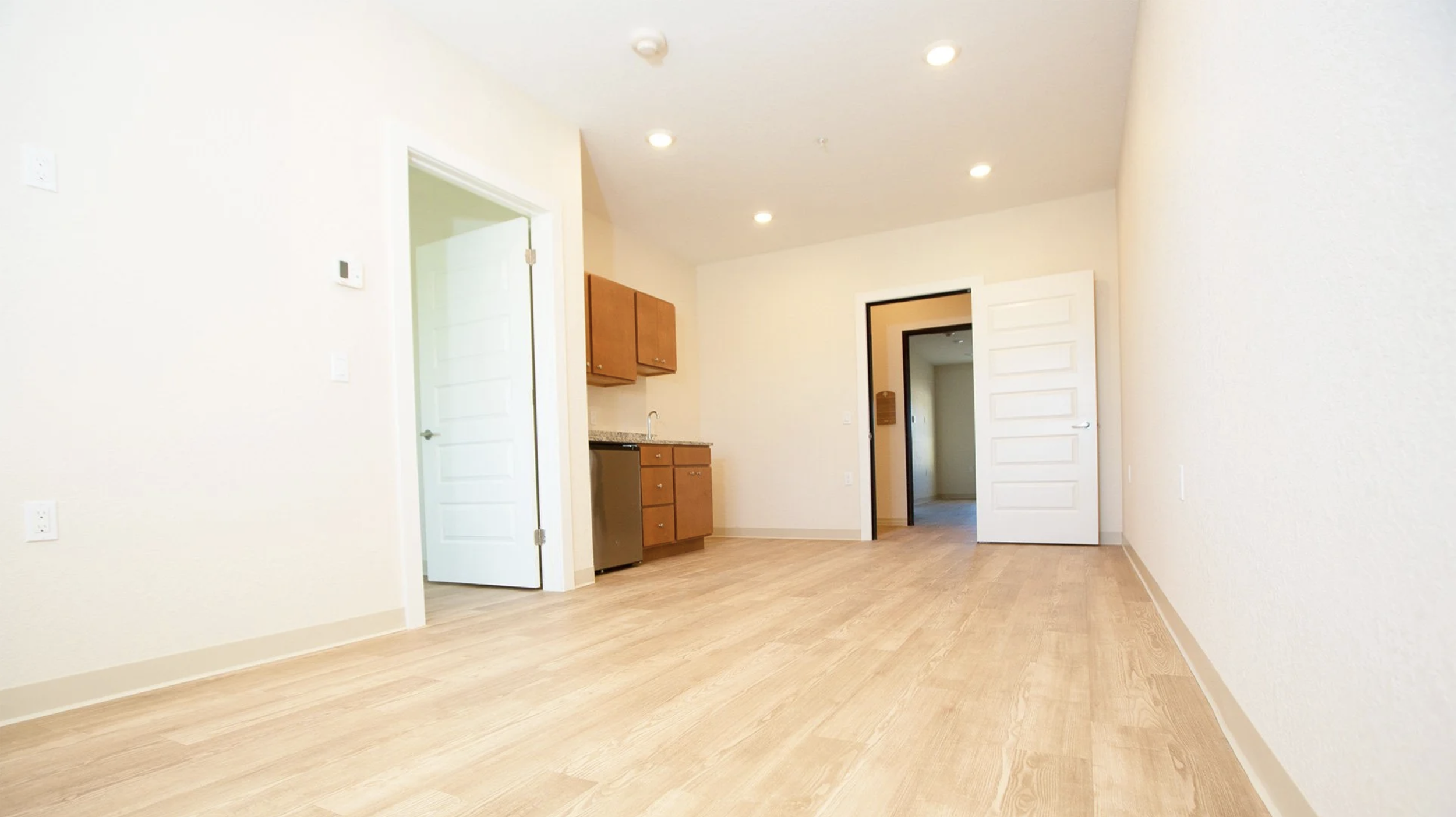 Empty room with light-colored walls and wood flooring, featuring a small kitchenette with wooden cabinets and a compact fridge, with multiple ceiling lights and open doors leading to other rooms.