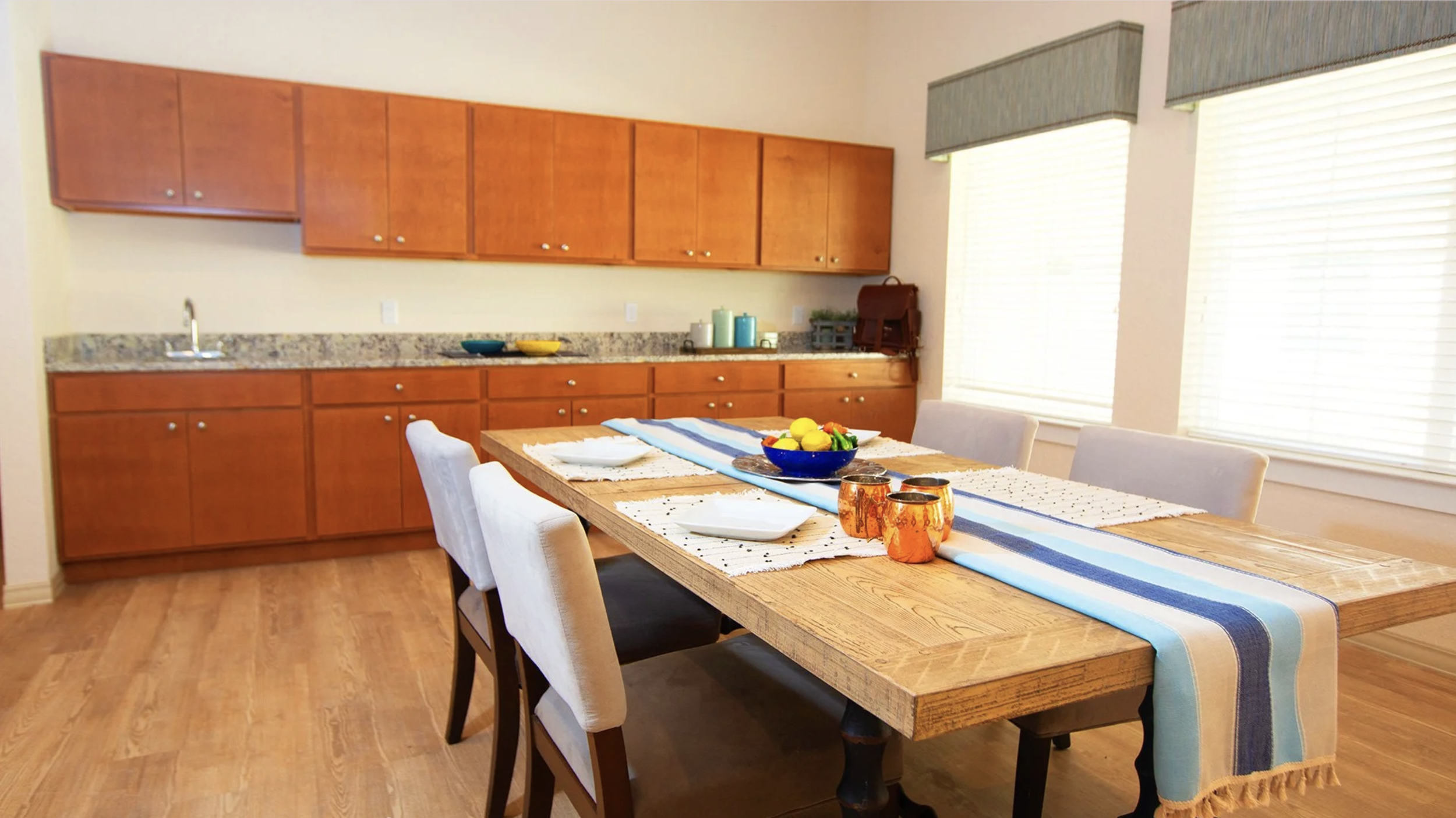 Dining room with wooden table, picnic-style bench, and four beige upholstered chairs. Table is decorated with a striped table runner, a bowl of lemons, and candles. Kitchen with wooden cabinets, granite countertop, and windows with blinds visible in 