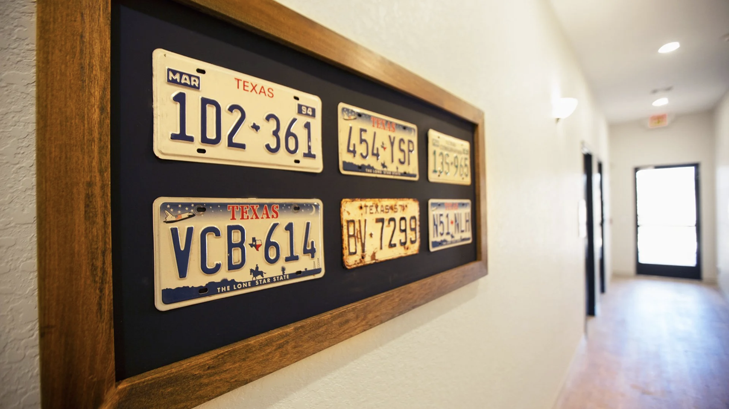 A framed display of vintage Texas license plates on a black background, hung on a beige wall in a hallway.