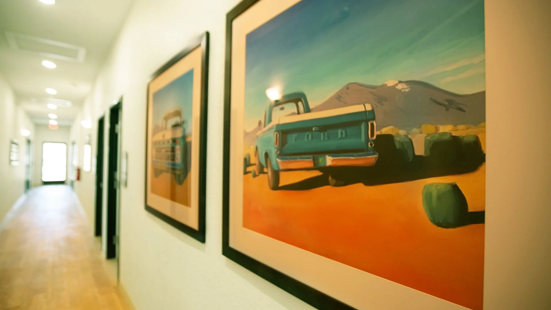 Hallway with framed landscape paintings, focusing on a painting of a vintage car against mountains and sky.