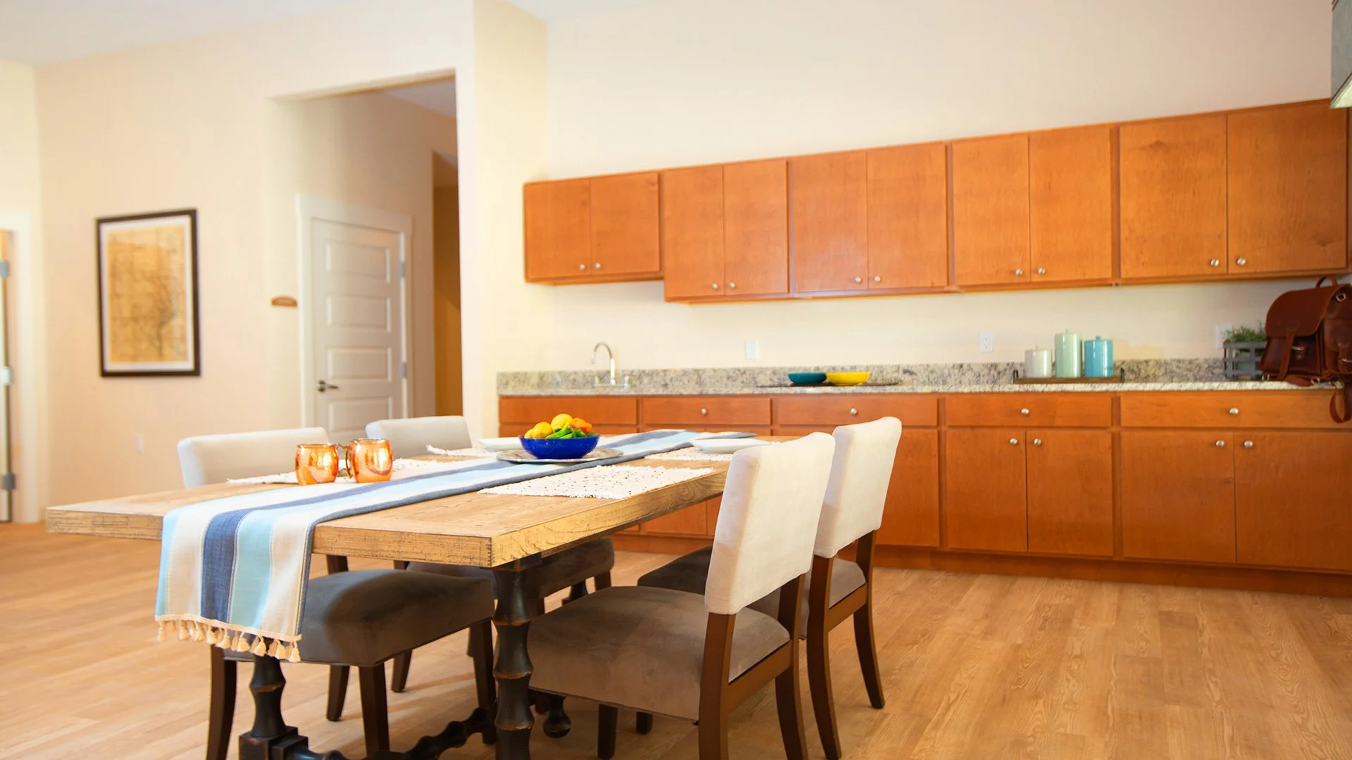 Dining area with a wooden table, six chairs, and a kitchen in the background with wooden cabinets and a granite countertop.