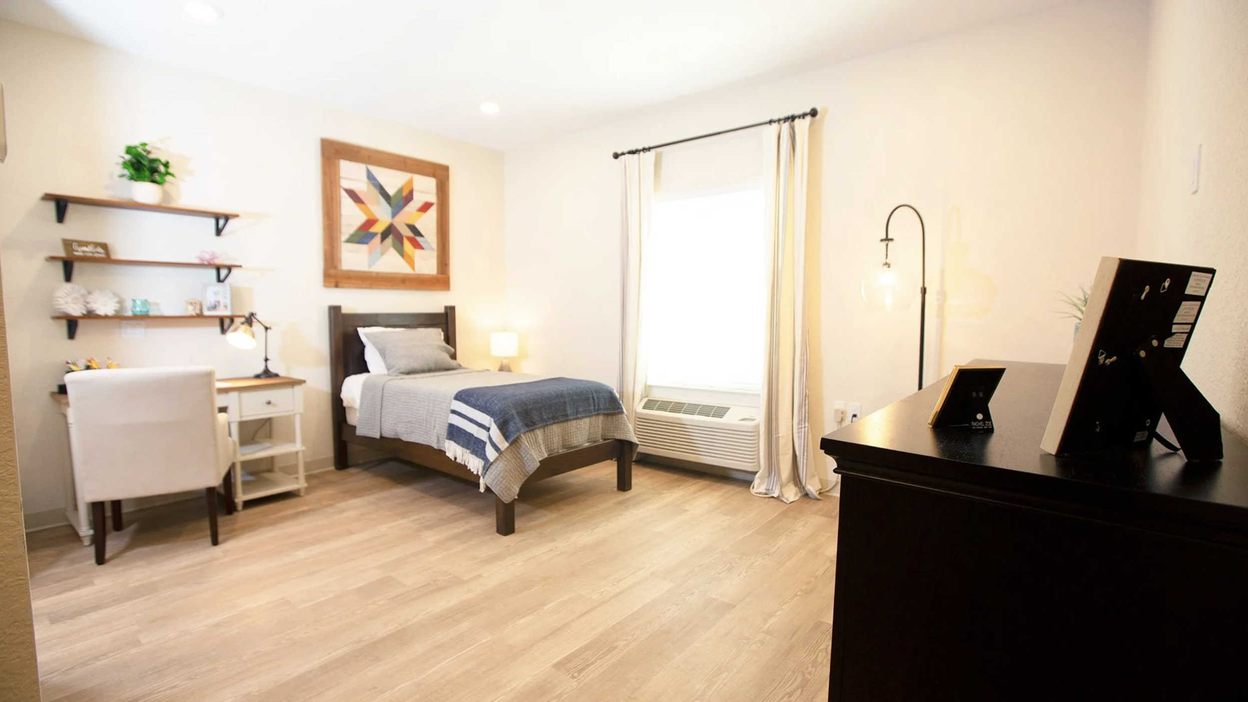A cozy bedroom with a bed, two pillows, a blanket, a nightstand with a lamp, wall shelves decorated with plants and pictures, a framed colorful geometric artwork, a chair, and a dresser with framed photos. The room is well-lit with natural light from