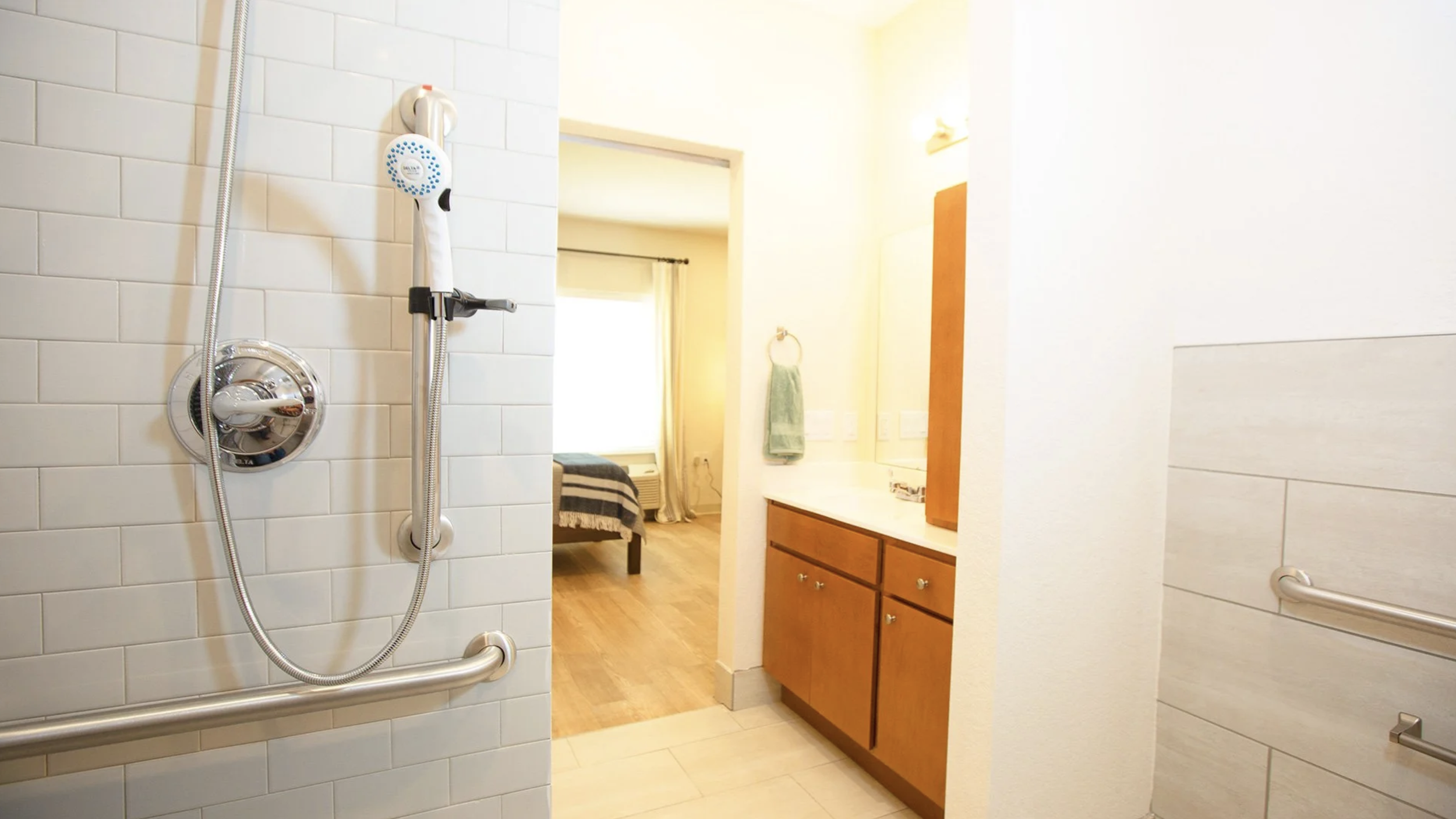 Bathroom with a walk-in shower, white tiled walls, metallic grab bars, a wooden vanity with a mirror, green towels, and a view into a bedroom with a bed and window.