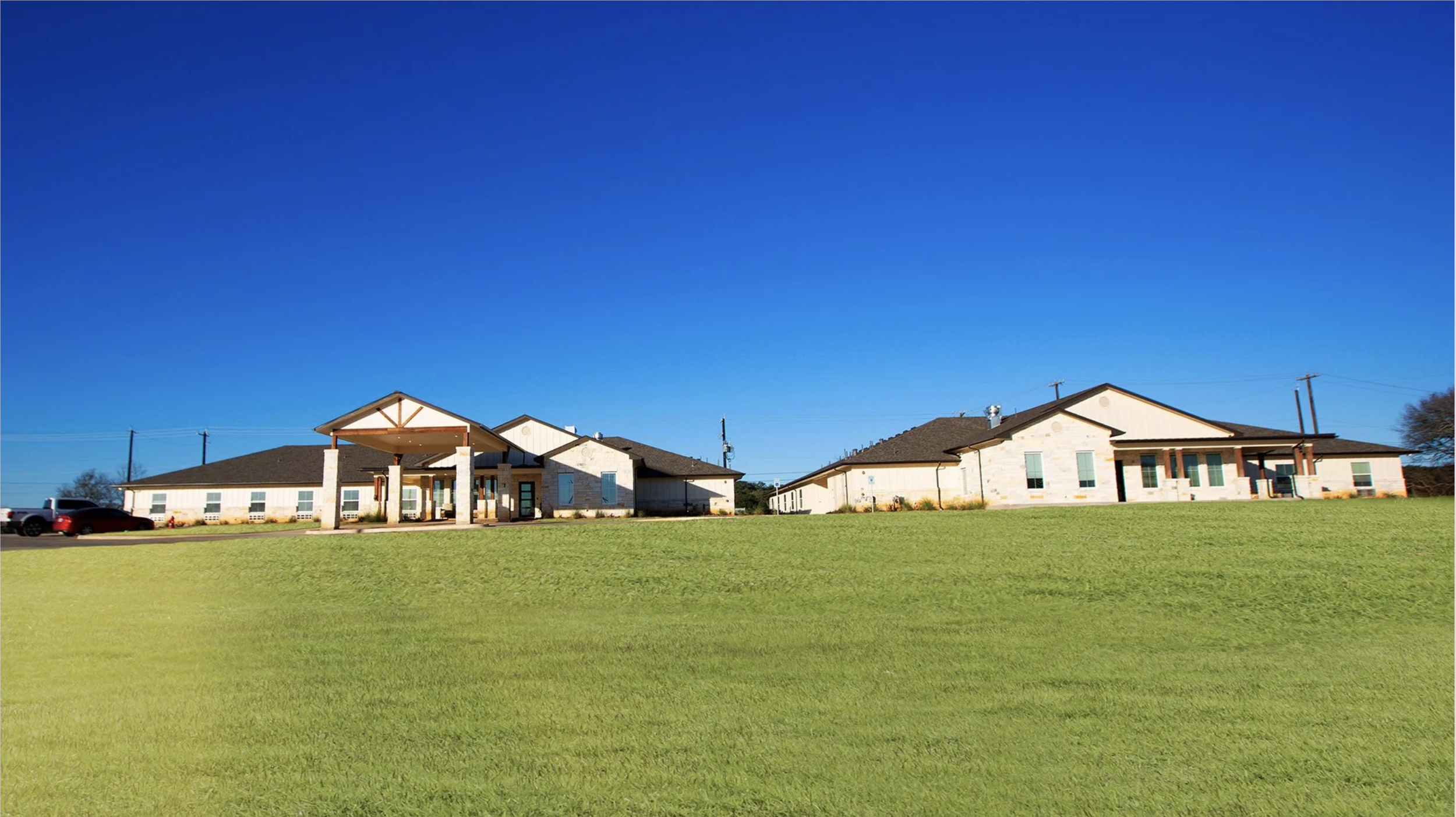 Modern single-story building with a stone exterior, large windows, and a covered entrance, set against a clear blue sky and green grass.
