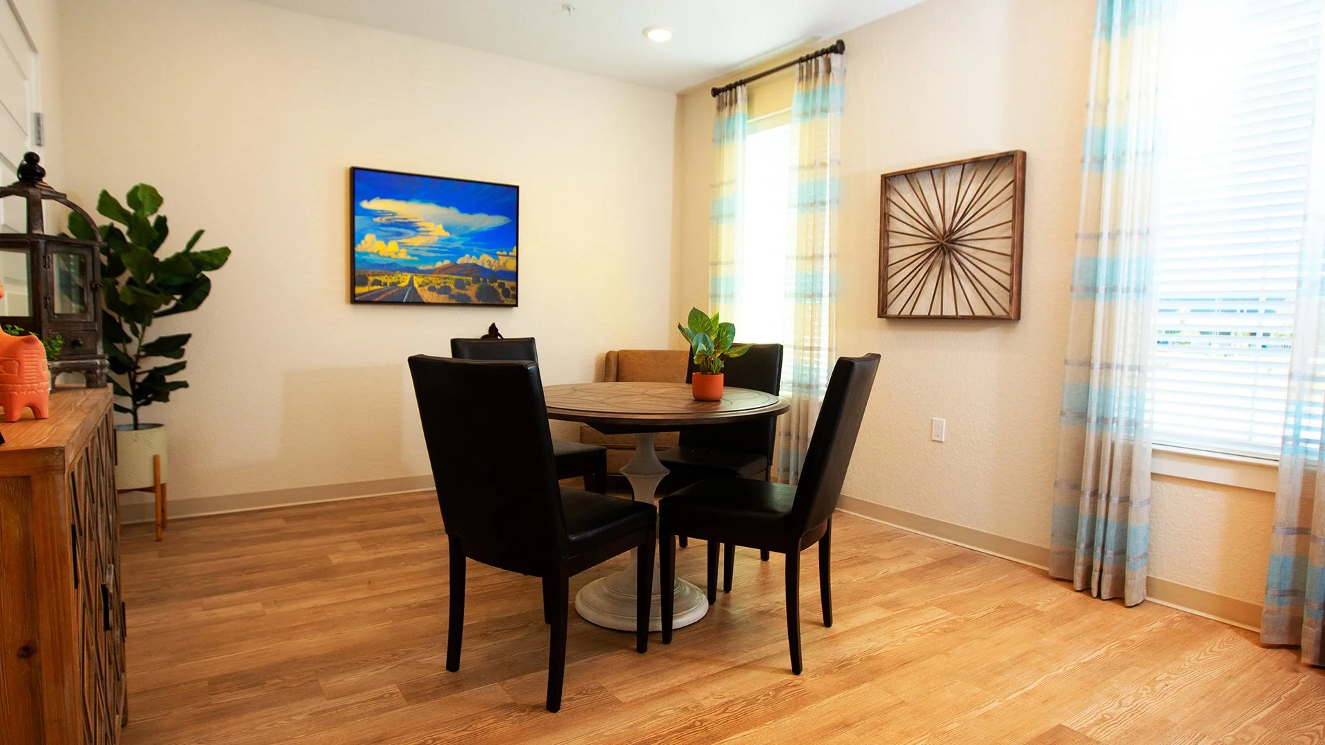 A cozy dining room with a round wooden table surrounded by four black chairs, a small potted plant in an orange pot on the table, a brown cushioned chair against the wall, a painting of a landscape with a road and mountains, and an abstract wooden wa
