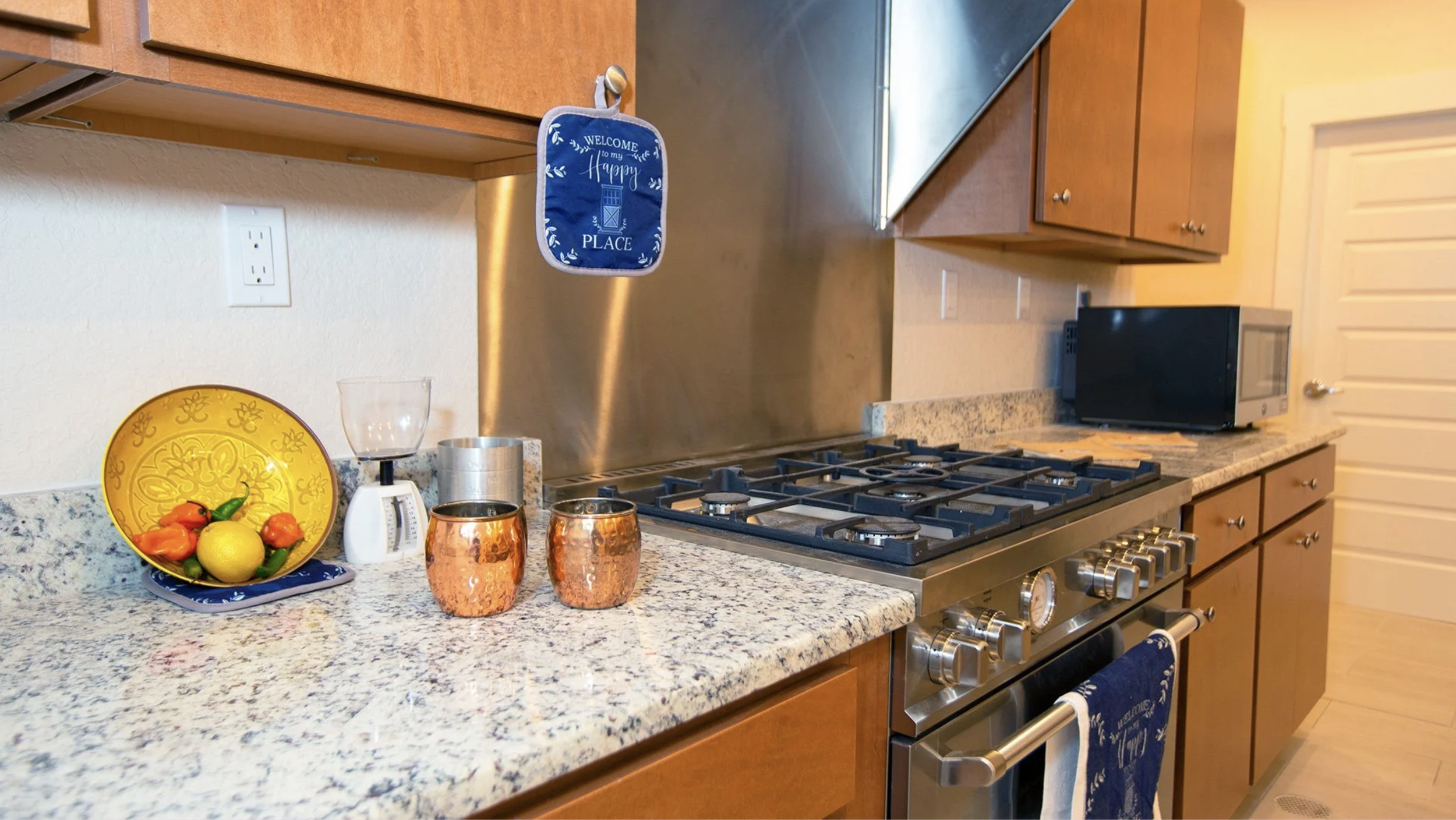Kitchen countertop with a bowl of fruit, copper cups, and kitchen appliances including a stove, microwave, and toaster oven, with wooden cabinets and a granite surface.