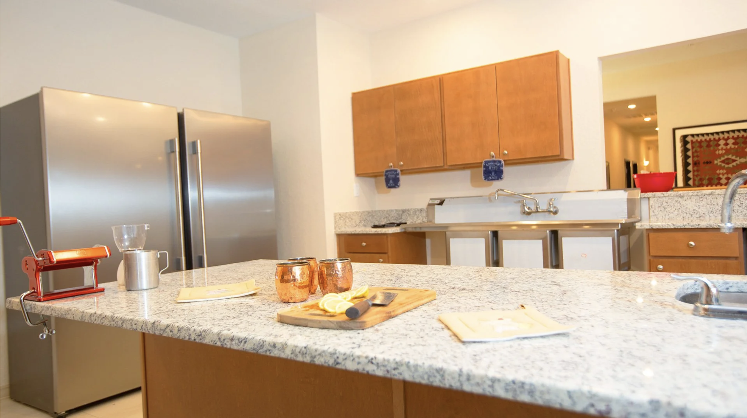 Kitchen with granite countertops, wooden cabinets, a stainless steel refrigerator, a double sink, and a kitchen island with lemons, copper mugs, and a pizza cutter. A red manual pasta machine is on the island.