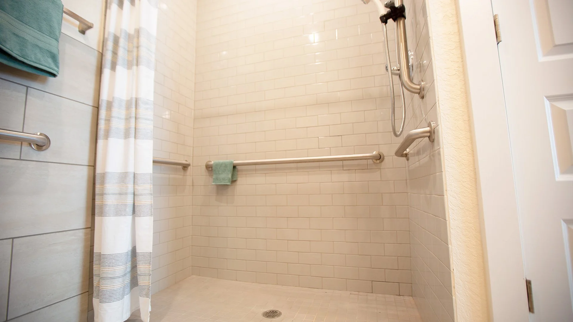 Accessible walk-in shower with white tile walls and beige floor, equipped with grab bars, a fold-down seat, and a handheld showerhead.