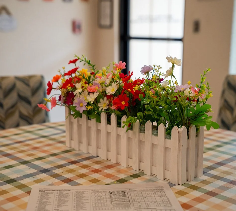 A white wooden planter box filled with colorful artificial flowers sits on a checkered tablecloth, with a window in the background.
