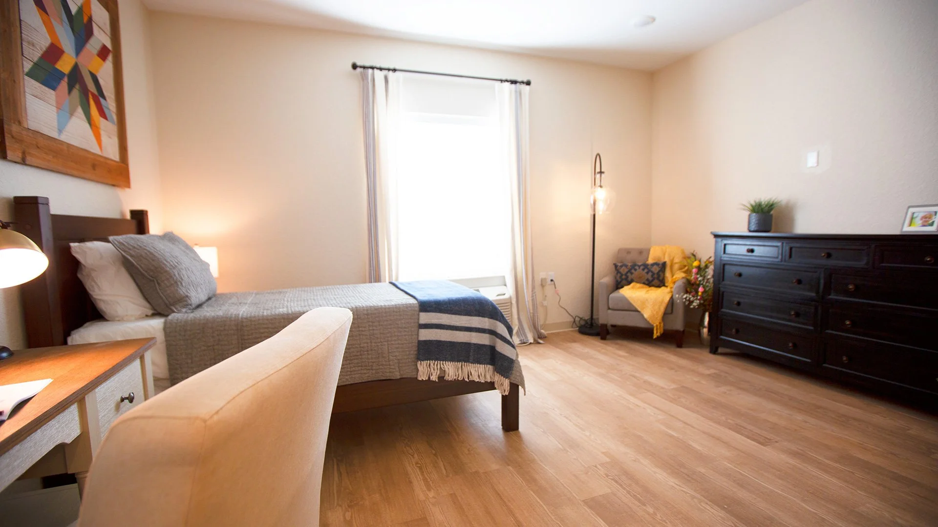 A bedroom with a bed, nightstand, armchair, dresser, and a window with curtains, decorated with plants and artwork.