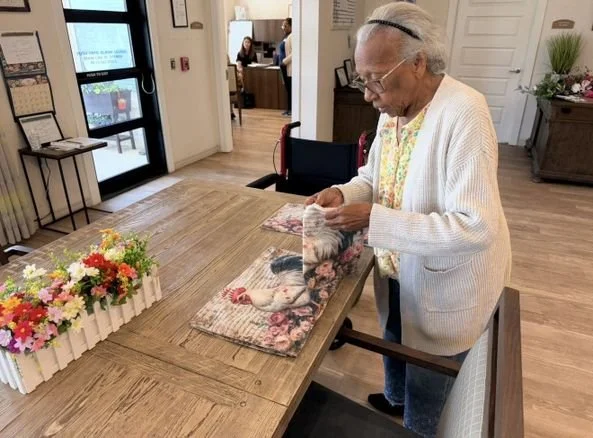 An elderly woman standing at a wooden table, holding a plastic bag of flowers or decorative items, inside a well-lit room with flowers on the table.