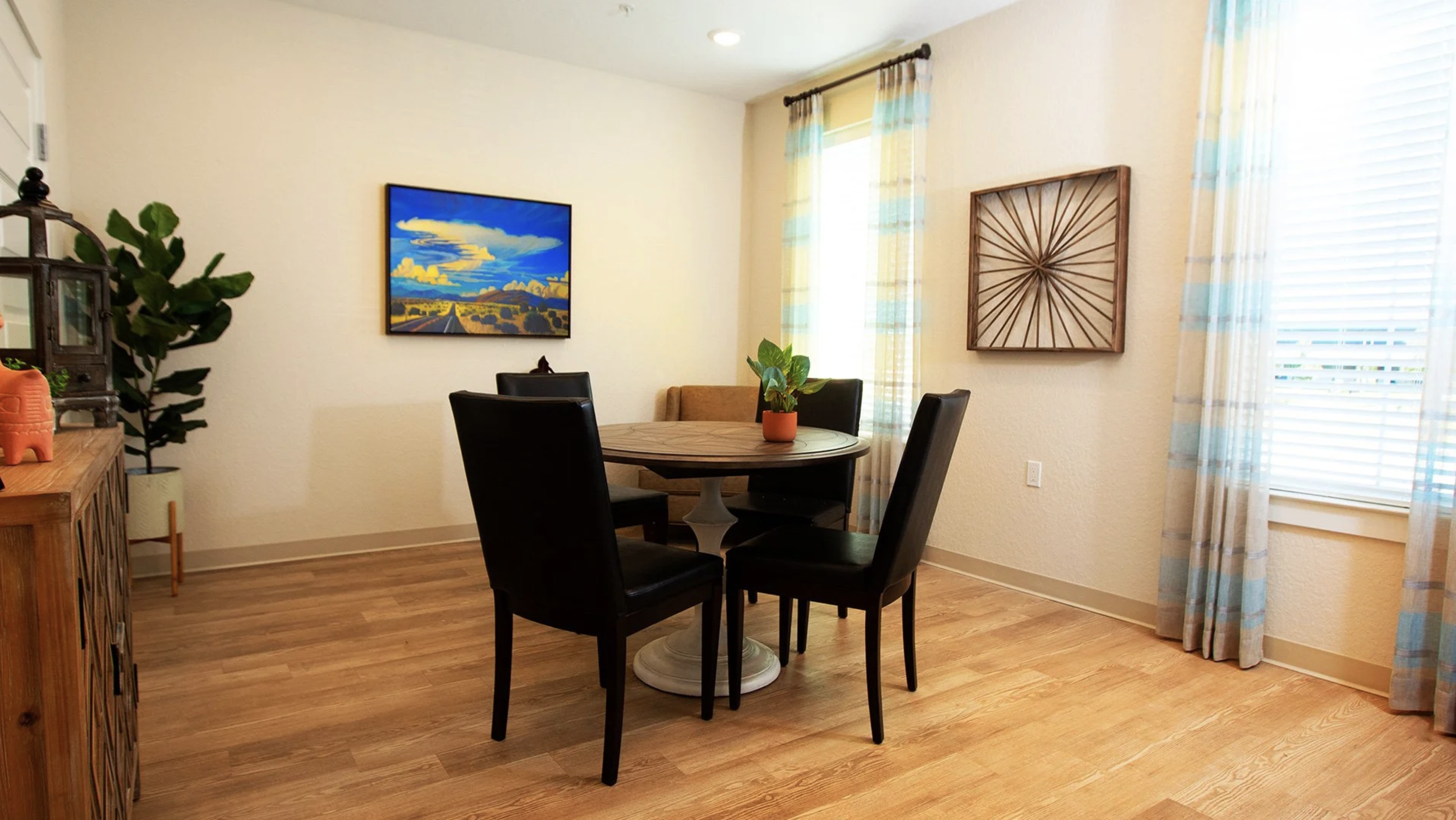 A cozy dining room with a round wooden table surrounded by four black chairs, a small potted plant in an orange pot on the table, a brown cushioned chair against the wall, a painting of a landscape with a road and mountains, and an abstract wooden wa