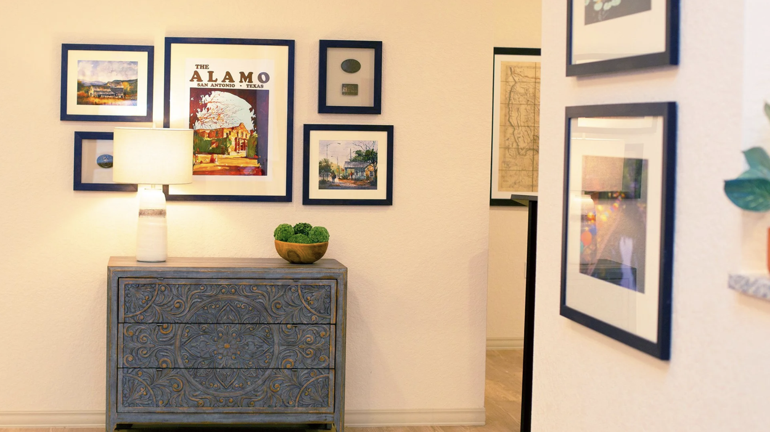 Interior wall with framed artwork, a decorative blue chest, a table lamp, and a bowl of green decorative orbs.