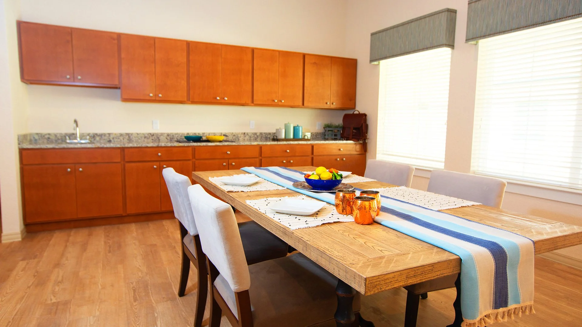 Dining room with wooden table, picnic-style bench, and four beige upholstered chairs. Table is decorated with a striped table runner, a bowl of lemons, and candles. Kitchen with wooden cabinets, granite countertop, and windows with blinds visible in 