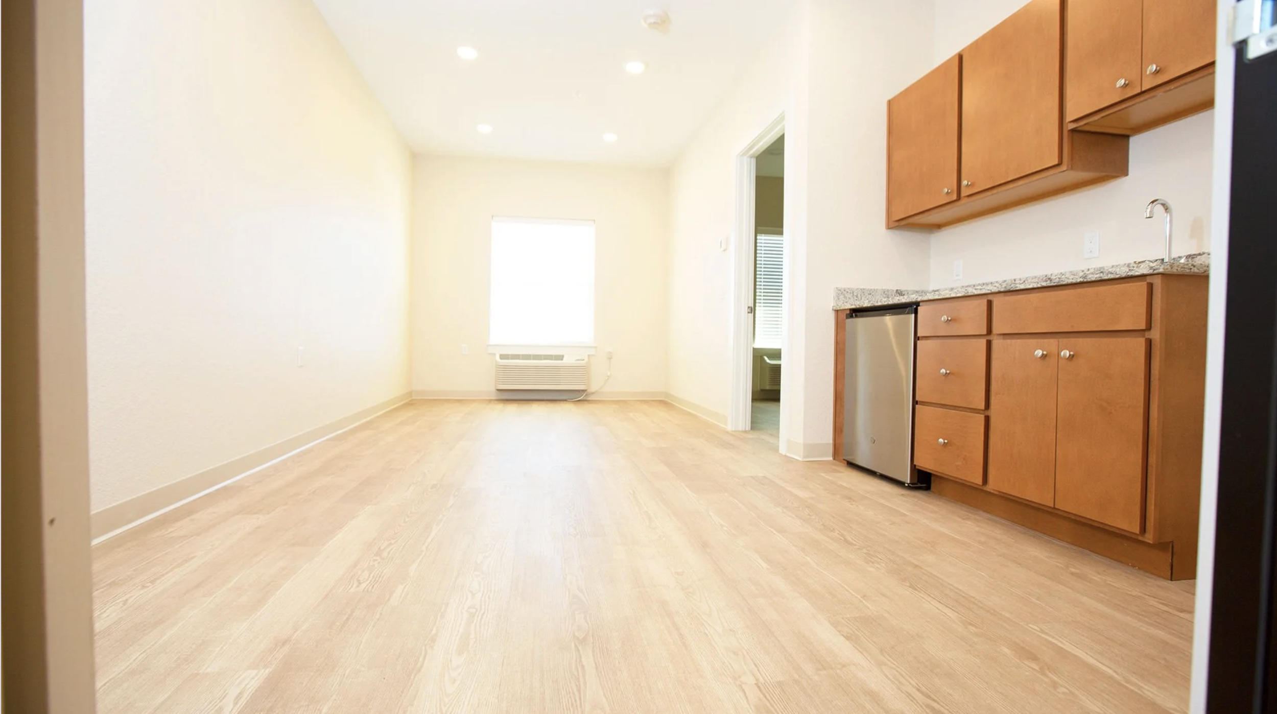 Empty room with light-colored wooden floors, beige walls, and large window with blinds. Kitchen area on the right side with brown cabinets, granite countertop, a small refrigerator, and a sink. Open doorway to another room.