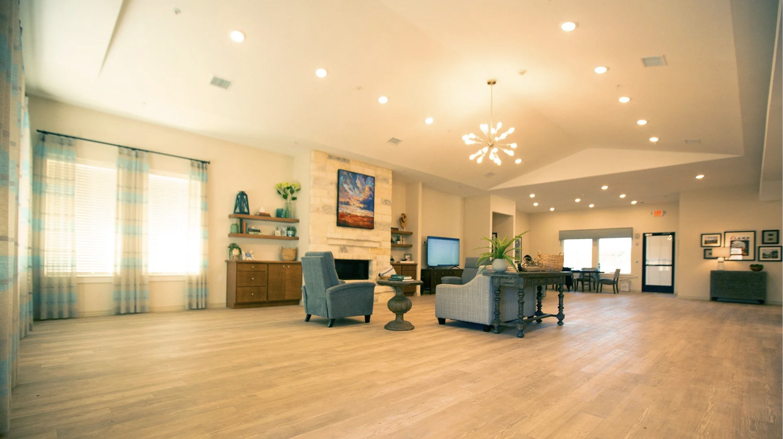 Spacious living room with beige walls, multiple windows with light blue and yellow striped curtains, a stone fireplace with a TV mounted above, various type of chairs, a wooden coffee table with books, a vase with greenery, and a modern chandelier on