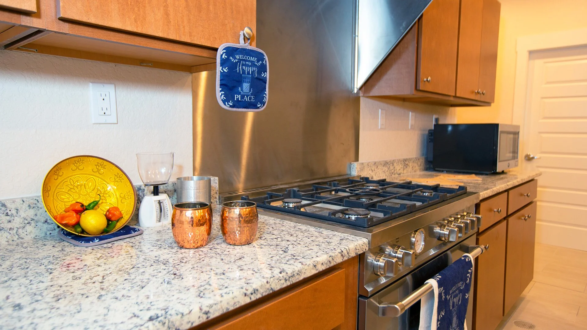 Kitchen countertop with a bowl of fruit, copper cups, and kitchen appliances including a stove, microwave, and toaster oven, with wooden cabinets and a granite surface.