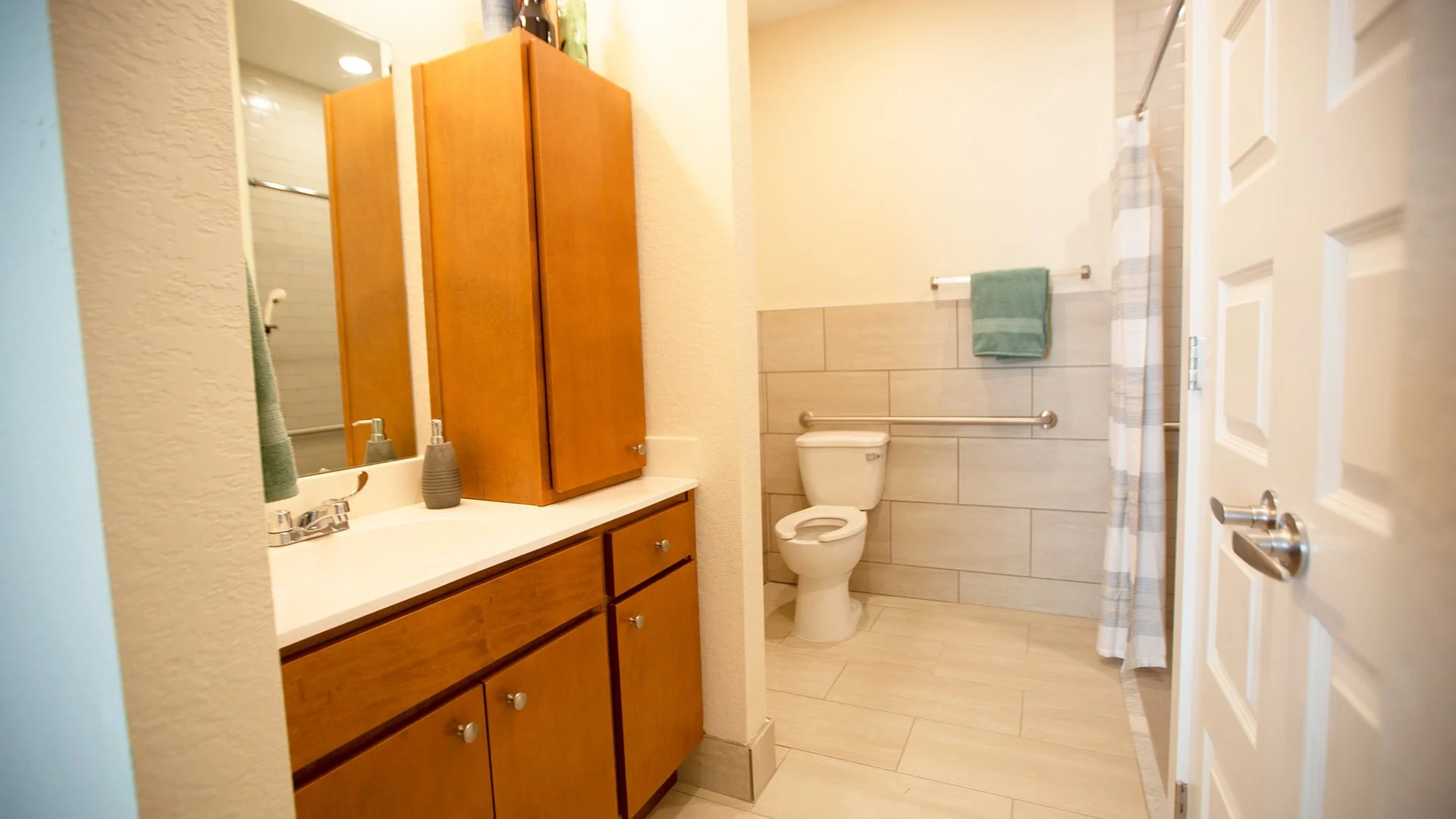 A bathroom with a wooden vanity, a mirror, a soap dispenser, a toilet, a towel bar with a green towel, a shower area with a striped curtain, and a door to the bathroom.