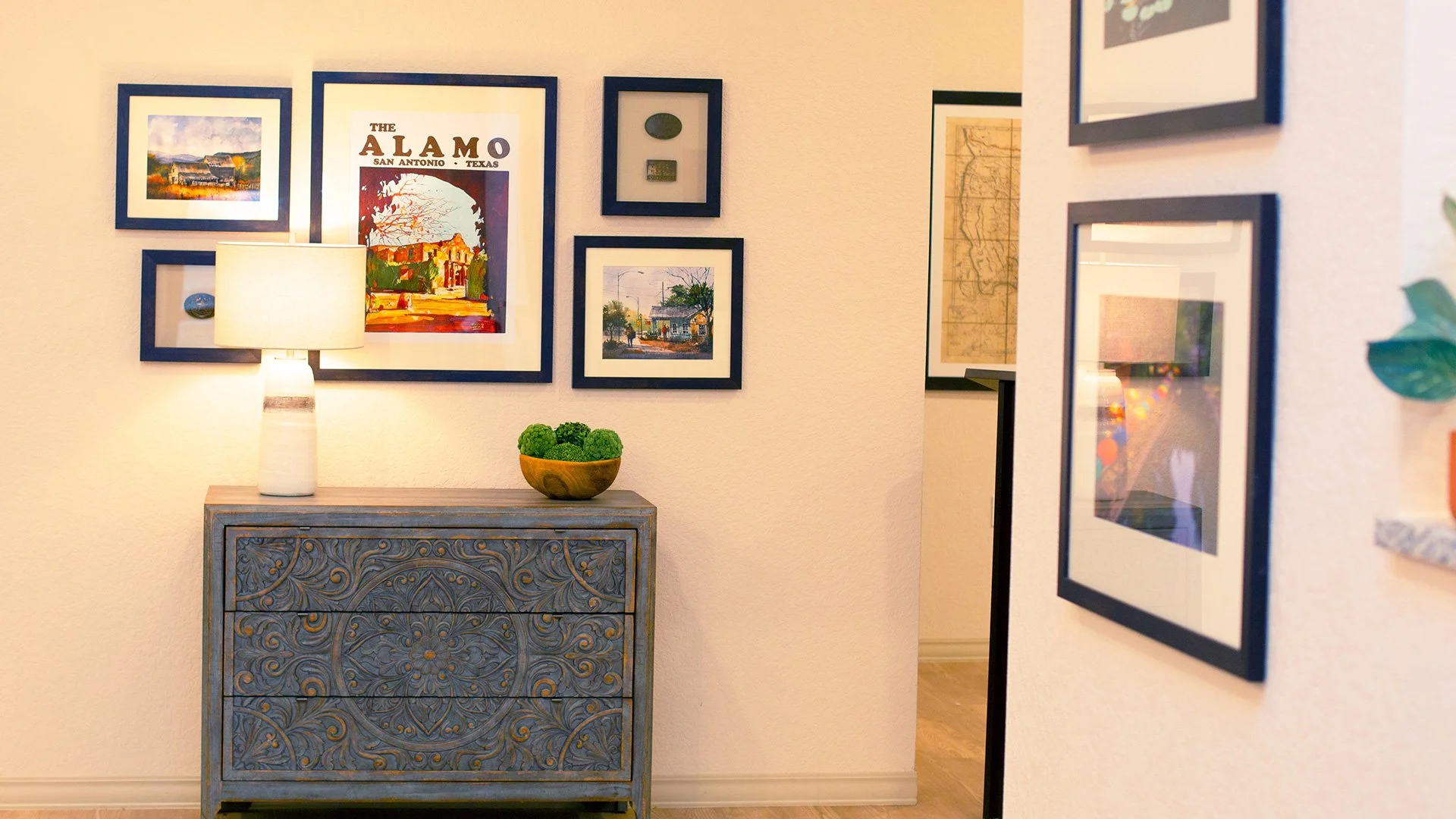Interior wall with framed artwork, a decorative blue chest, a table lamp, and a bowl of green decorative orbs.