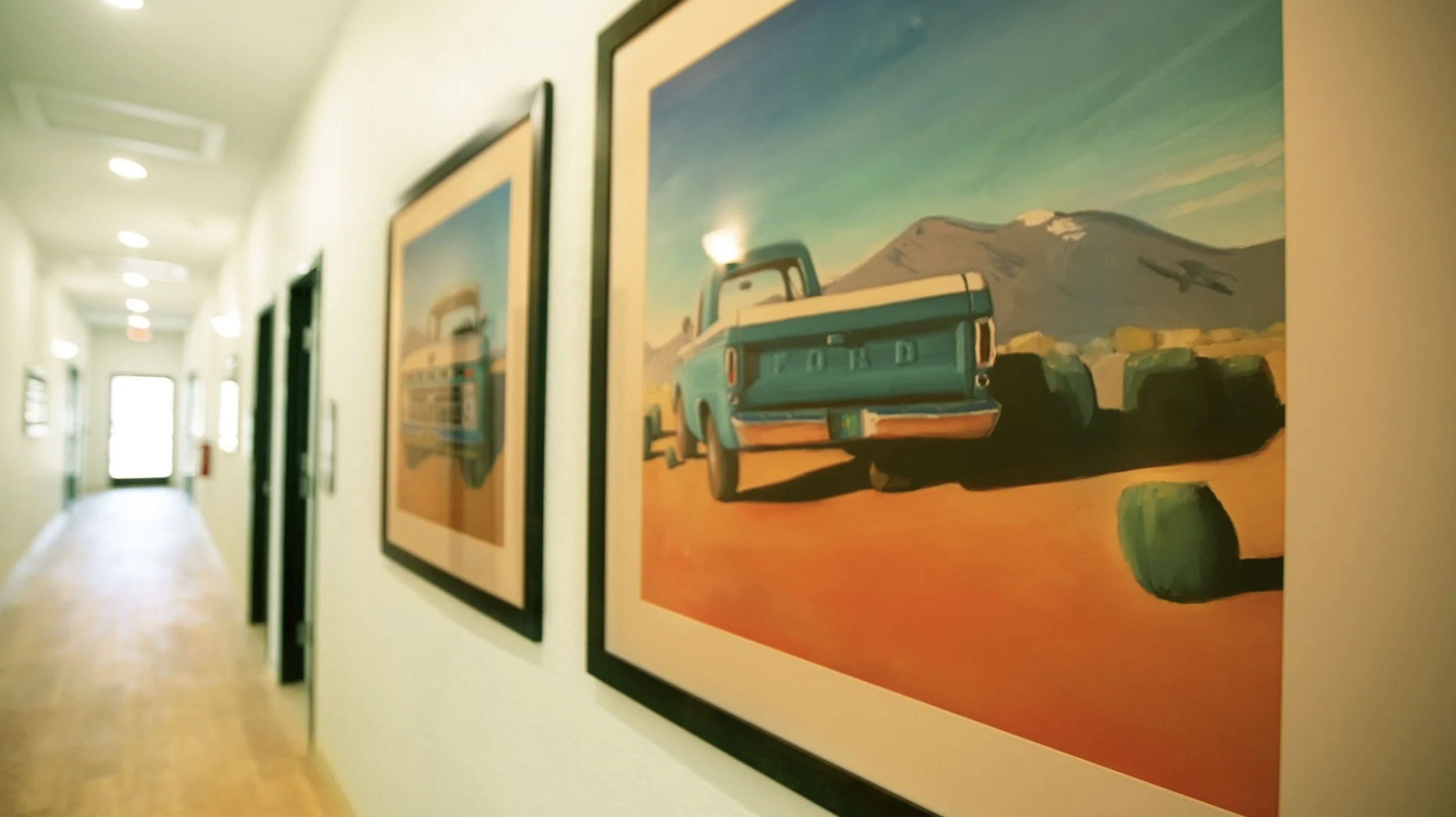 Hallway with framed landscape paintings, focusing on a painting of a vintage car against mountains and sky.