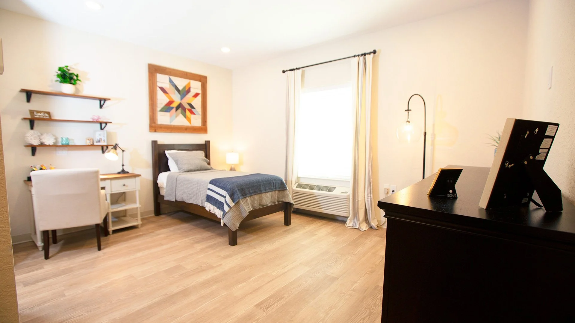 A cozy bedroom with a bed, two pillows, a blanket, a nightstand with a lamp, wall shelves decorated with plants and pictures, a framed colorful geometric artwork, a chair, and a dresser with framed photos. The room is well-lit with natural light from