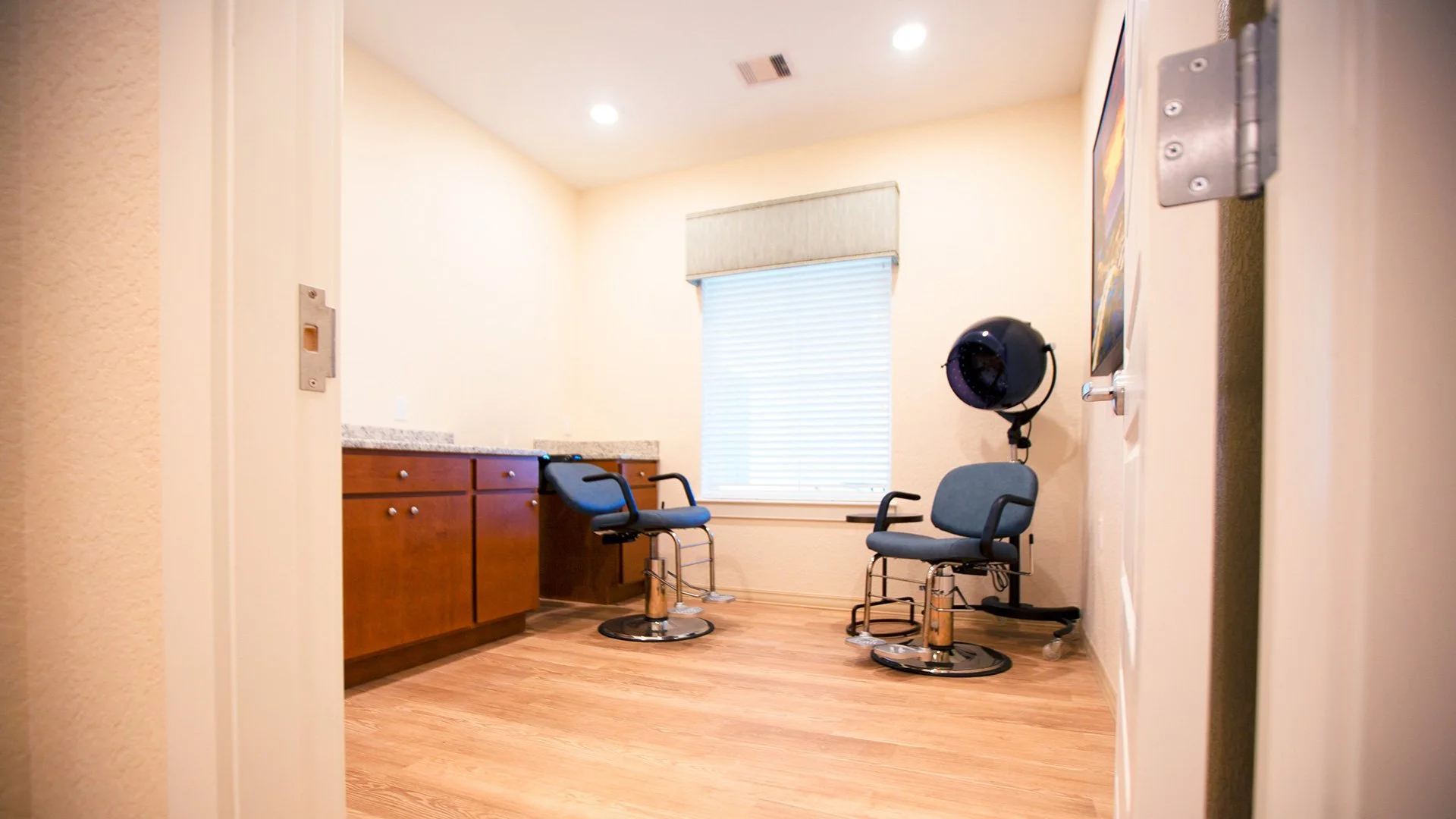 Room with two chairs, a hair dryer, cabinet, window with blinds, and wall art, likely a salon or barber shop waiting area.