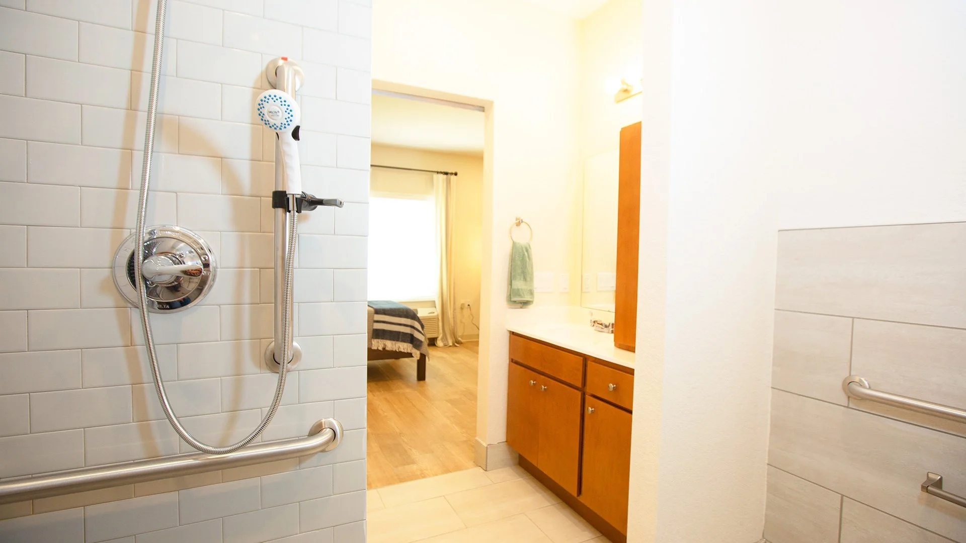 Bathroom with a walk-in shower, white tiled walls, metallic grab bars, a wooden vanity with a mirror, green towels, and a view into a bedroom with a bed and window.