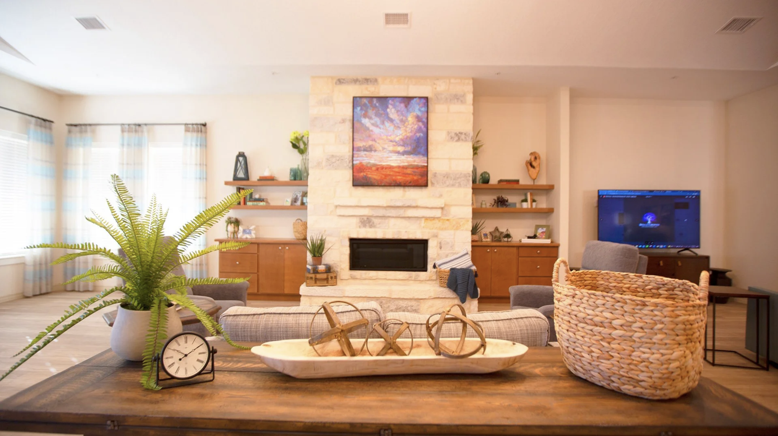 Living room with a central stone fireplace, some wall-mounted wooden shelves, a television, and a variety of decorative items. Potted plants and a dining table with a clock and decorative objects in the foreground.