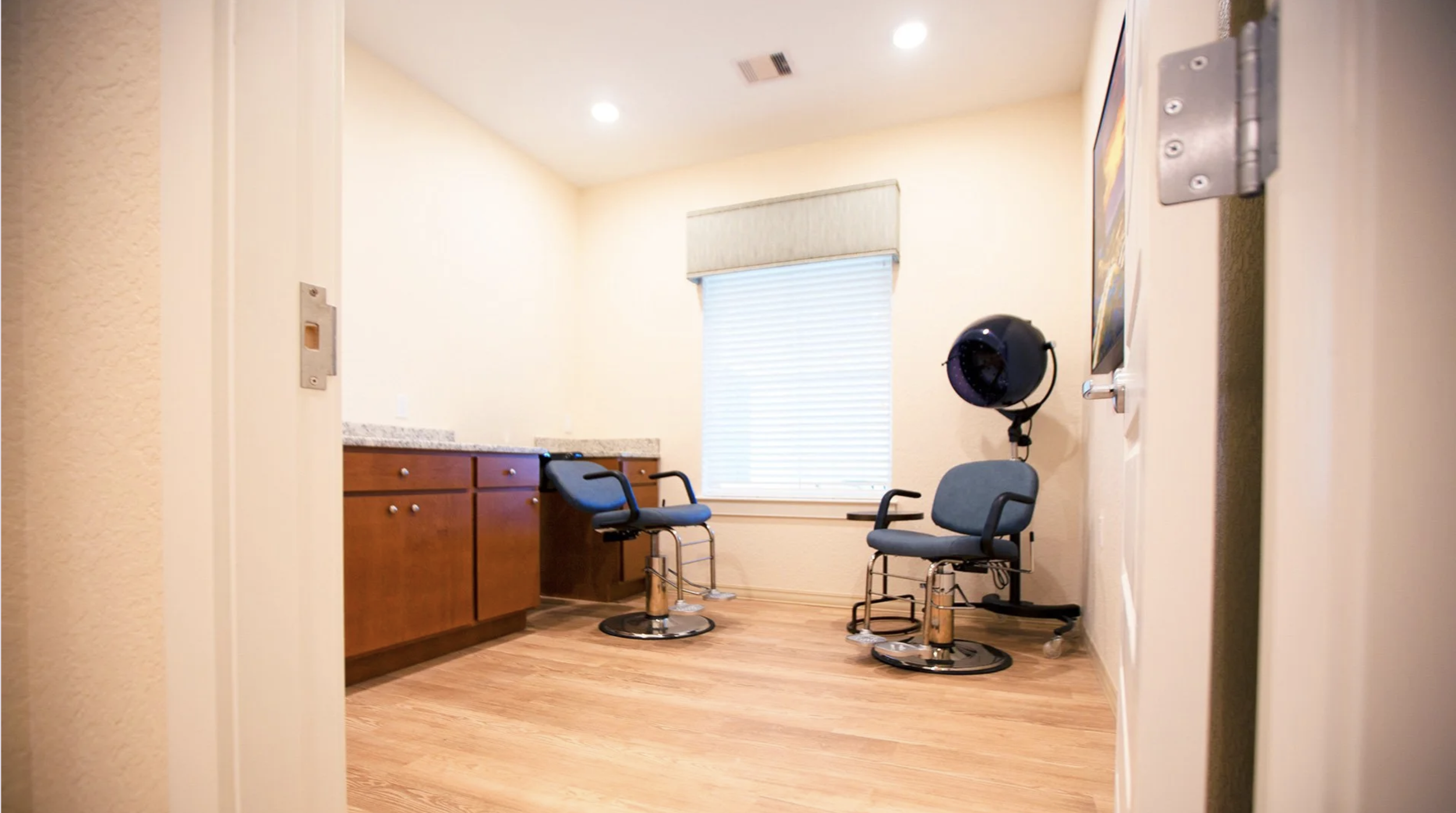 Room with two chairs, a hair dryer, cabinet, window with blinds, and wall art, likely a salon or barber shop waiting area.