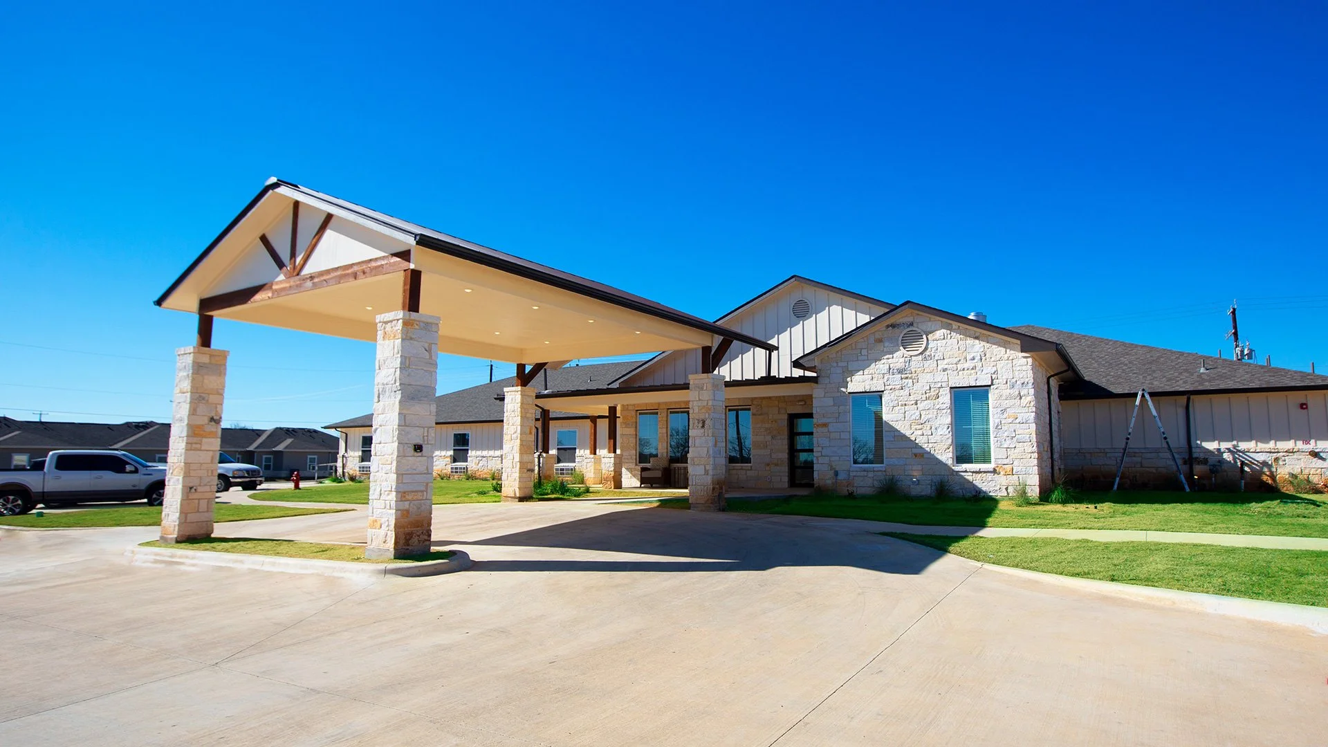 Modern residential building with stone and siding exterior, covered entrance with stone columns, parked vehicles, and clear blue sky.