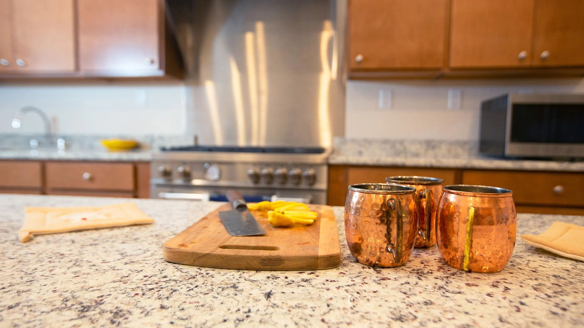 Kitchen countertop with three copper mugs, a wooden cutting board with sliced yellow peppers, a knife, a folded cloth, and a napkin in the foreground; kitchen cabinets, stove, microwave, and a silver backsplash in the background.