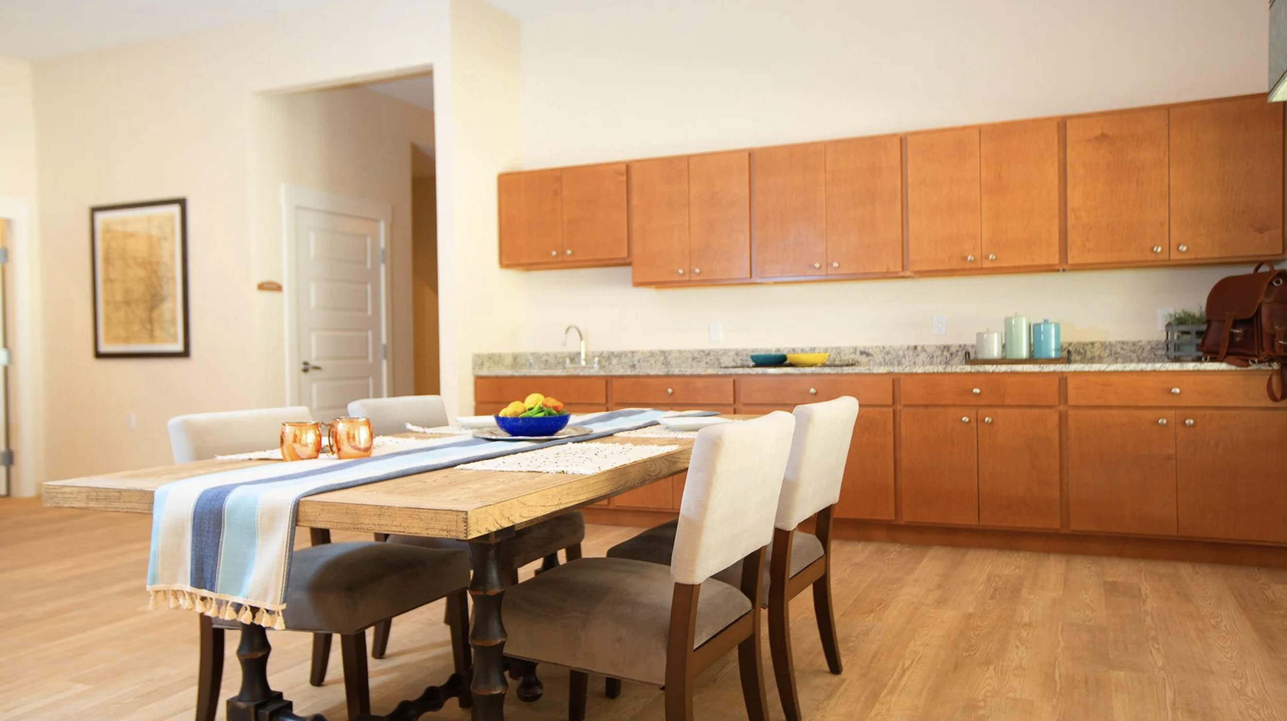 Dining area with a wooden table, six chairs, and a kitchen in the background with wooden cabinets and a granite countertop.