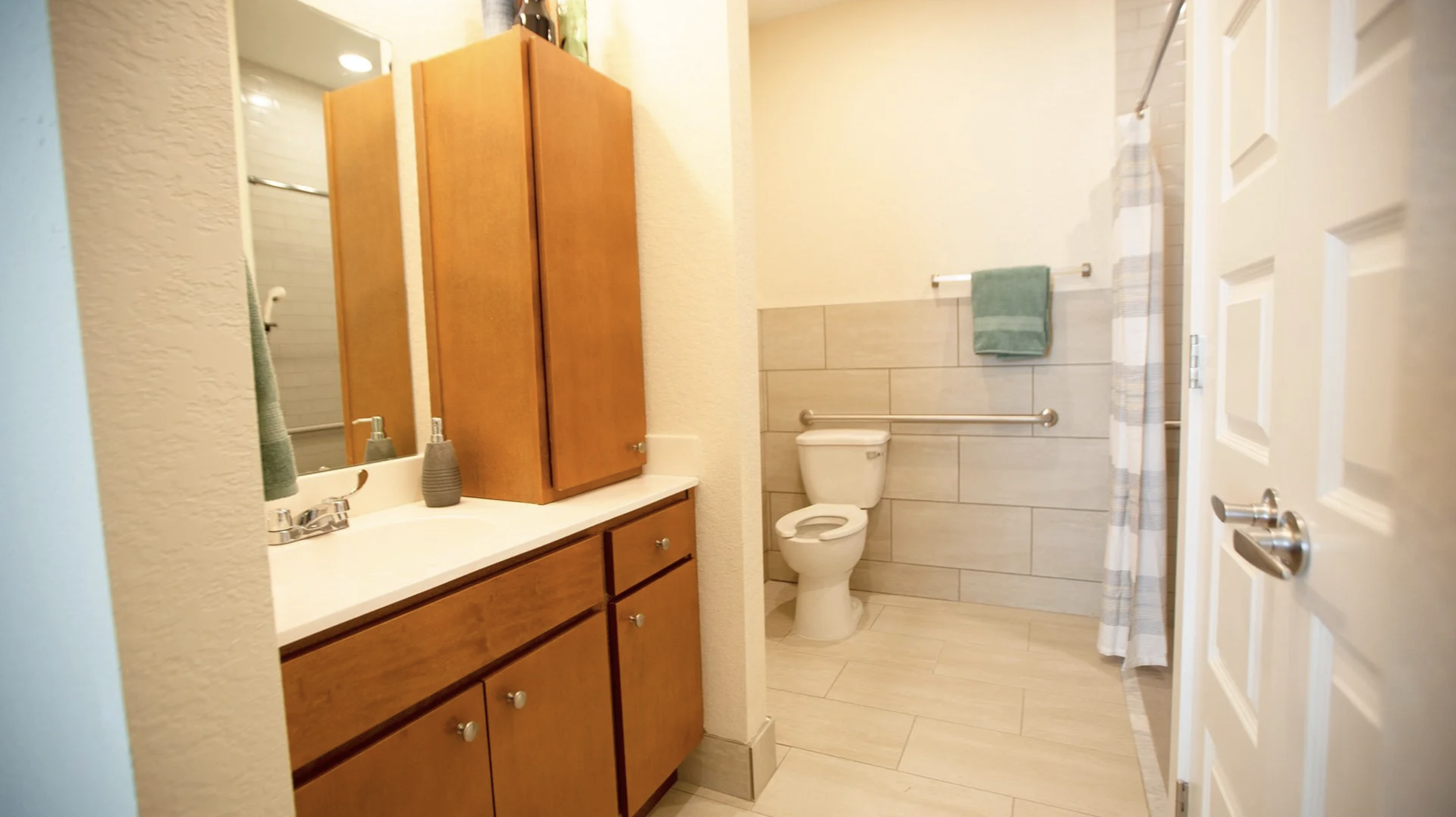 A bathroom with a wooden vanity, a mirror, a soap dispenser, a toilet, a towel bar with a green towel, a shower area with a striped curtain, and a door to the bathroom.