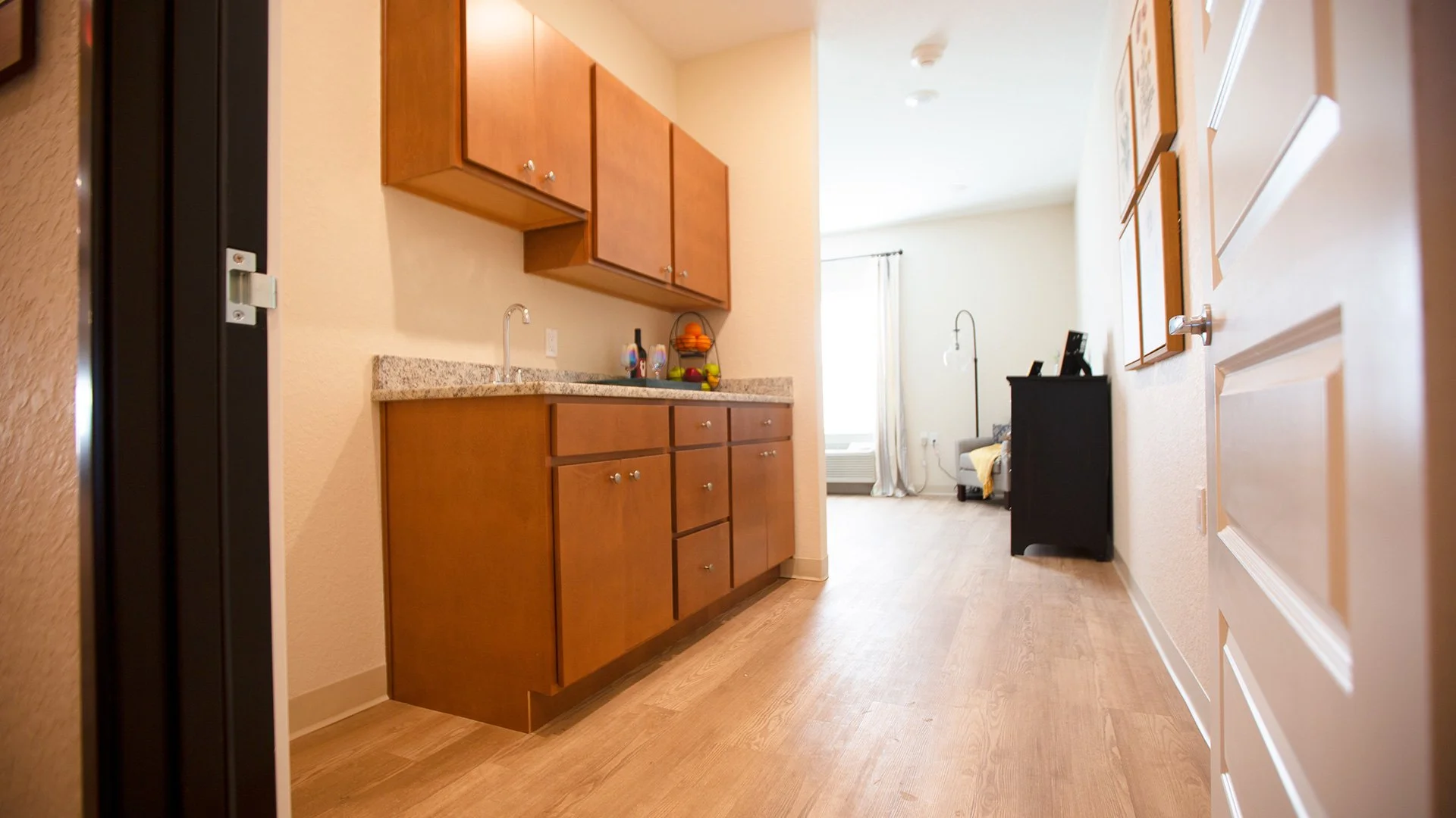 Interior view of a small apartment with a kitchen area featuring wooden cabinets, a granite countertop, and a sink. Past the kitchen, a living area is visible with a window, a radiator, and some furniture, including a black dresser or cabinet and a s